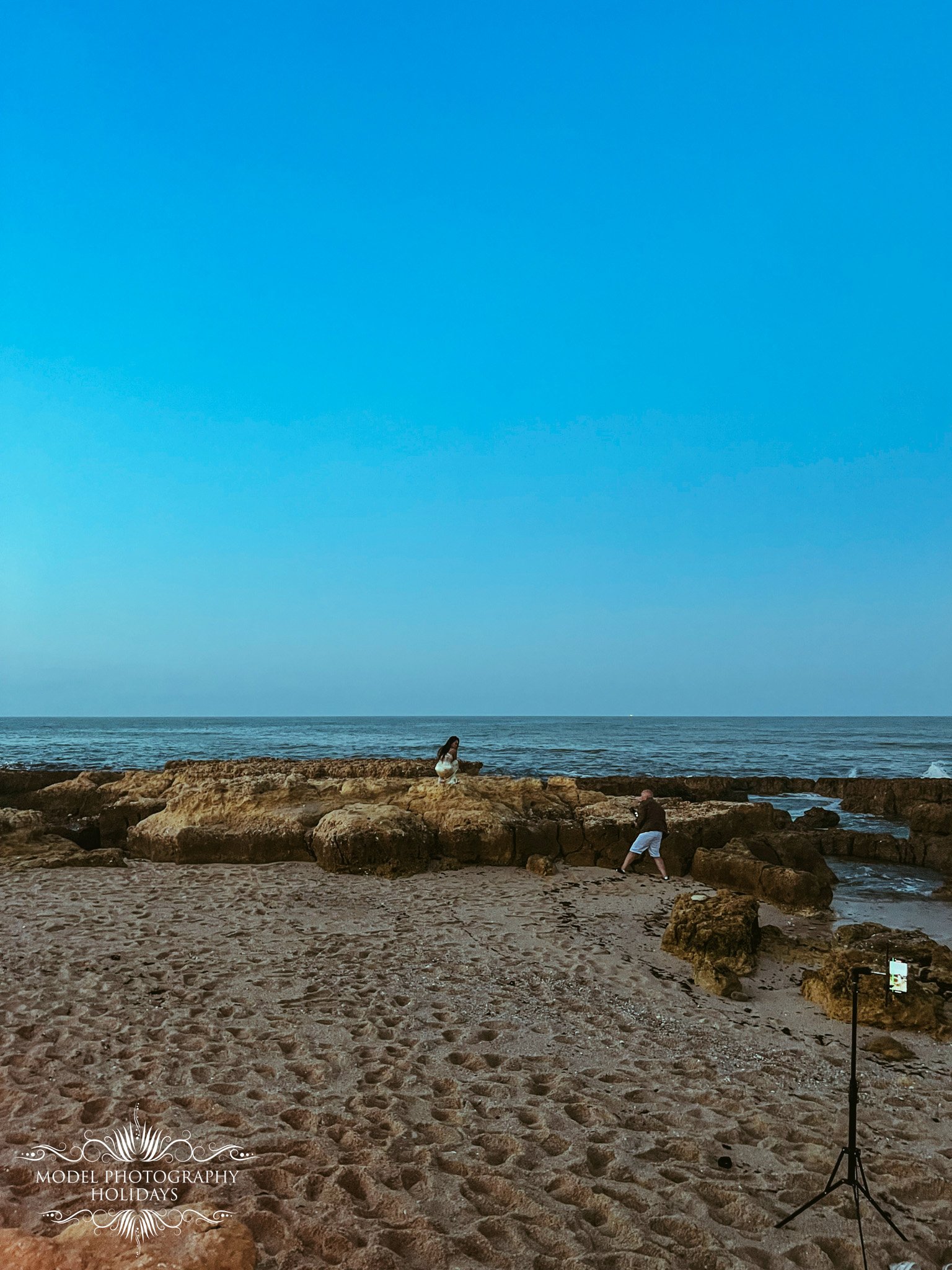 A beach scene with sand, rocks, and the ocean in the background. Two people, one sitting on rocks near the water and another standing further back, are present. A photography lighting stand is set up on the right side. The sky is blue with some cloud
