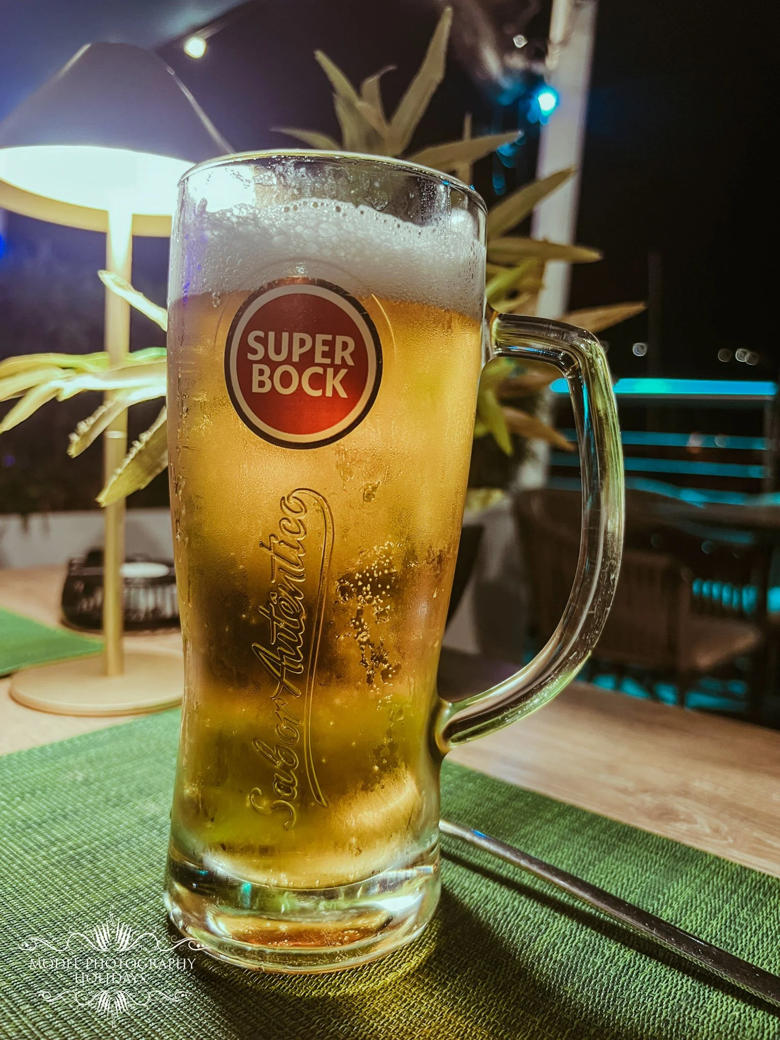 A glass mug filled with light-colored beer with foam on top, featuring a red and white 'Super Bock' logo, placed on a green textured placemat on a wooden table with a chopstick beside it. Background includes a lamp, a decorative plant, and blurred ou