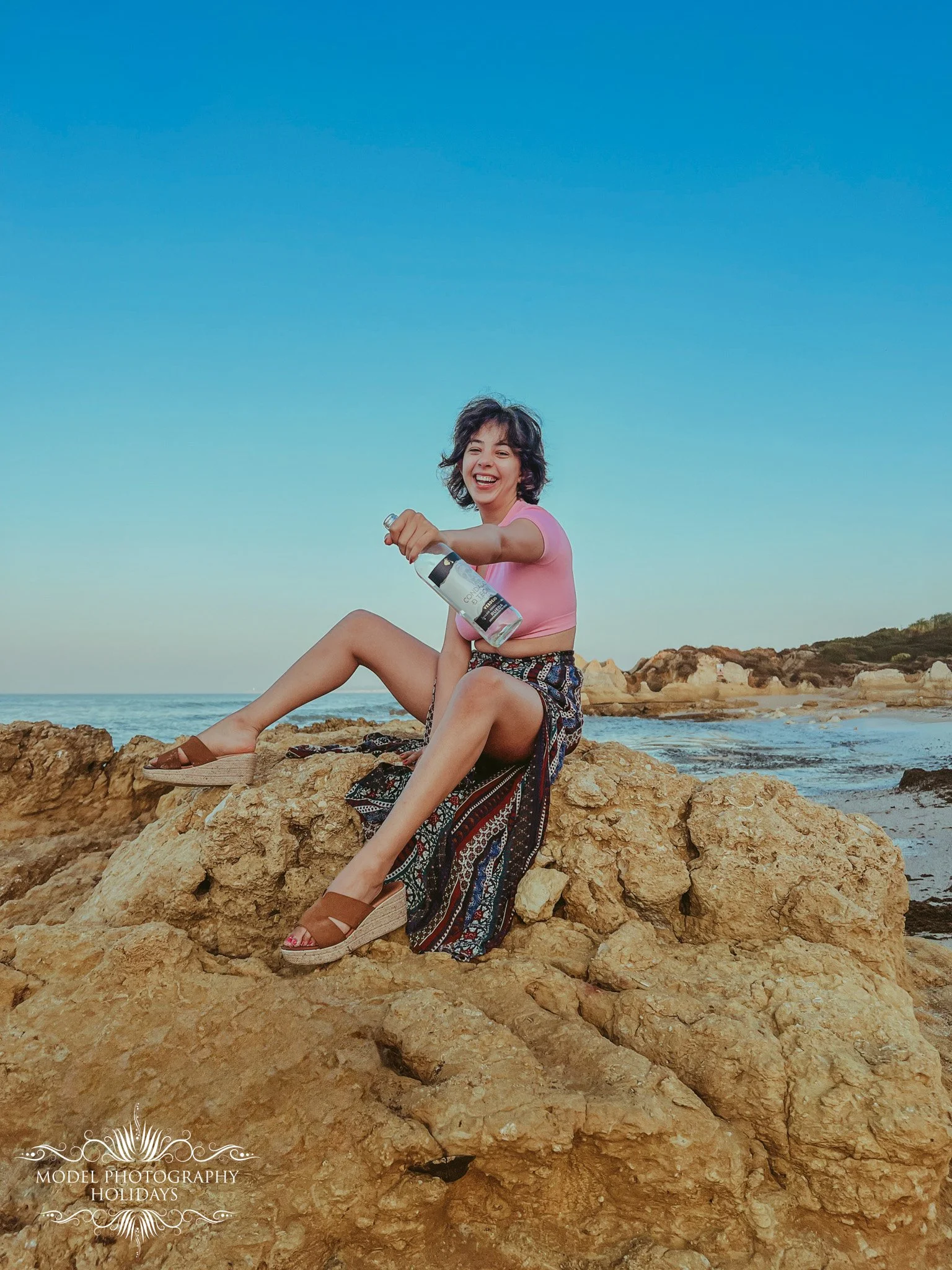 A woman sitting on rocks at the beach, smiling and holding a bottle of water, with the ocean and clear blue sky in the background.