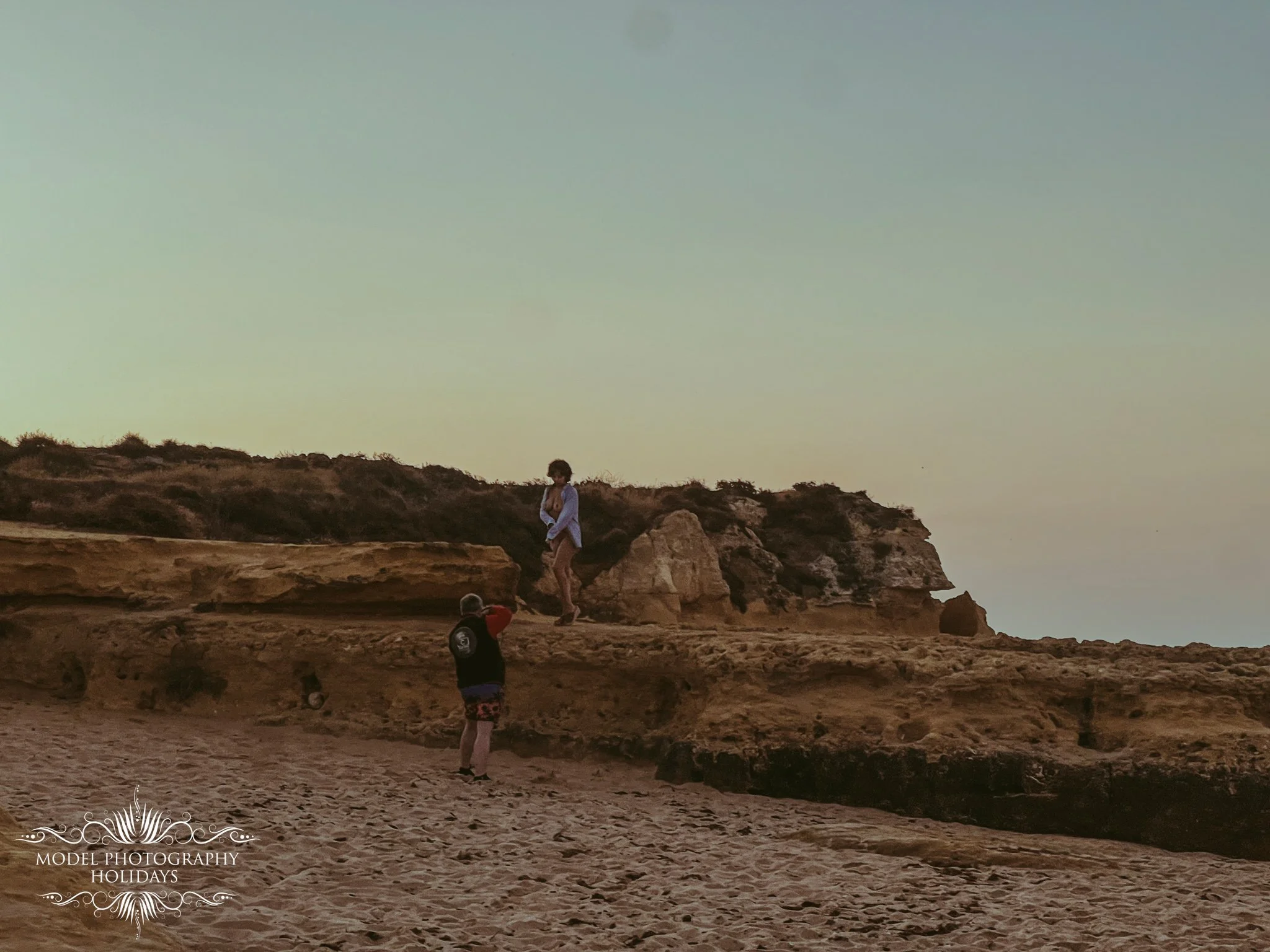 People enjoying a seaside cliff during sunset, with rocks and sandy beach in foreground, and a calm sky in the background.