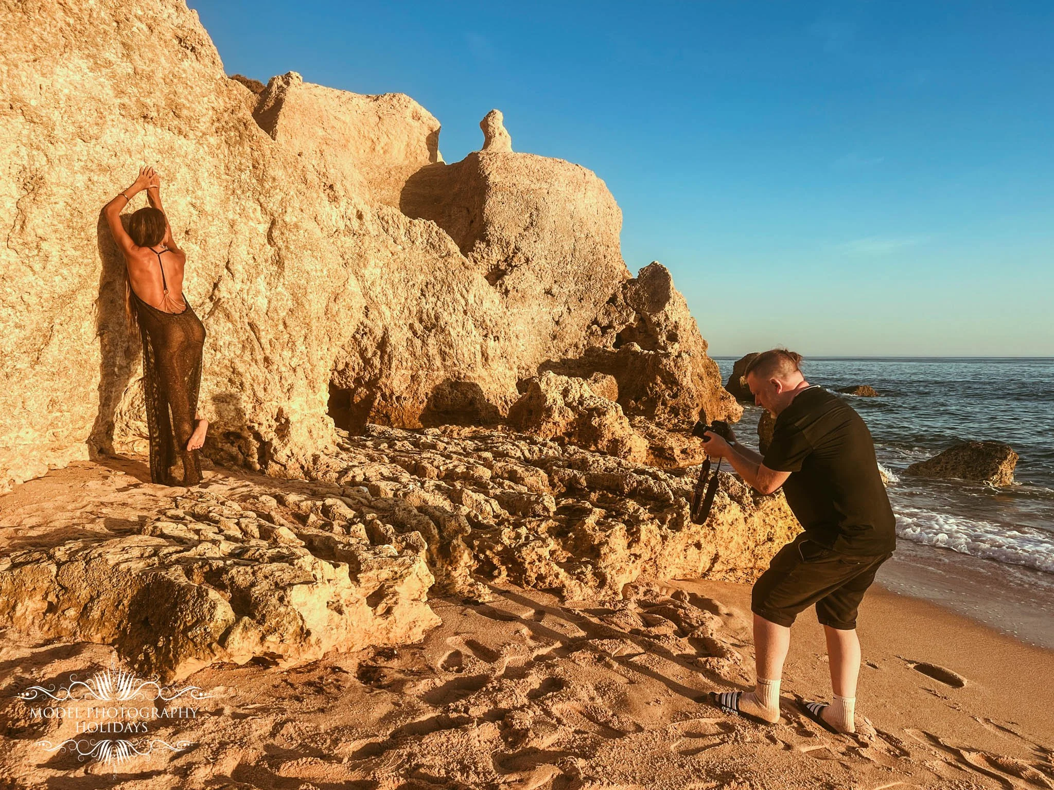 A model in a black, sheer dress stands on a rocky beach, with a large rock formation behind her and an ocean in the background. Two photographers are capturing her image, one is kneeling close to the ground while the other is standing and looking at 