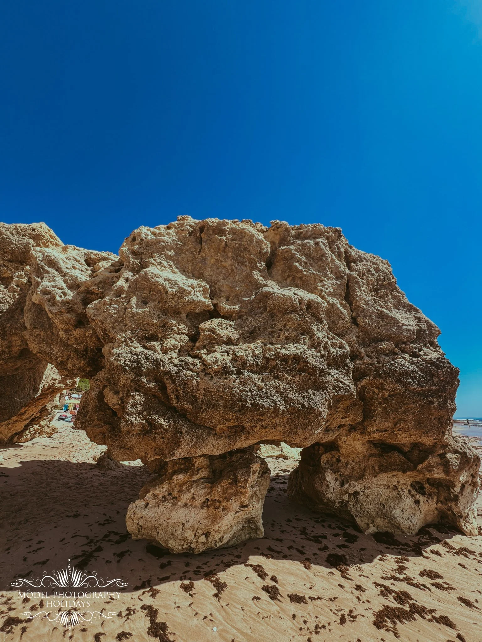 Large weathered rock formation on a sandy beach under a bright blue sky.