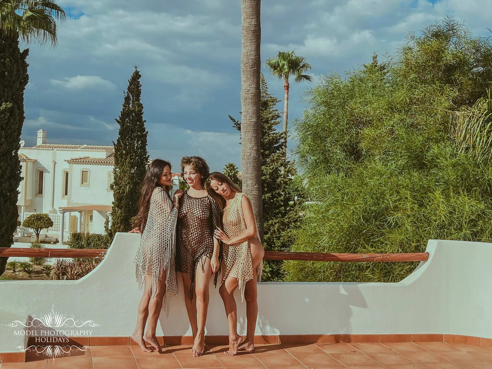Four women standing close together outdoors, dressed in lightweight, patterned dresses, smiling, with trees and a building with a white facade and red-tiled roof in the background.