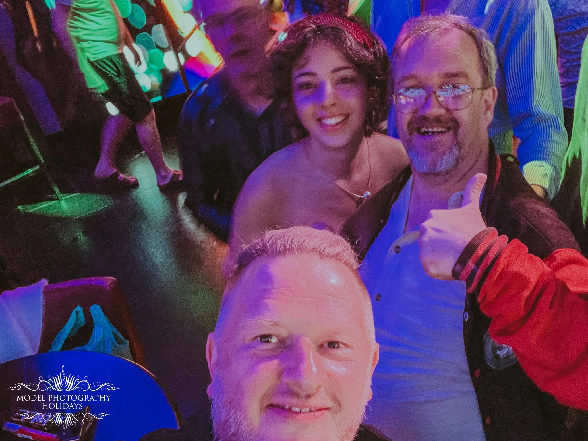 A group of people at a party or celebration with colorful lighting. Two people in the foreground are smiling, the woman has curly hair and the man has glasses and a beard. There is a logo in the bottom left corner that reads 'Model Photography Holida