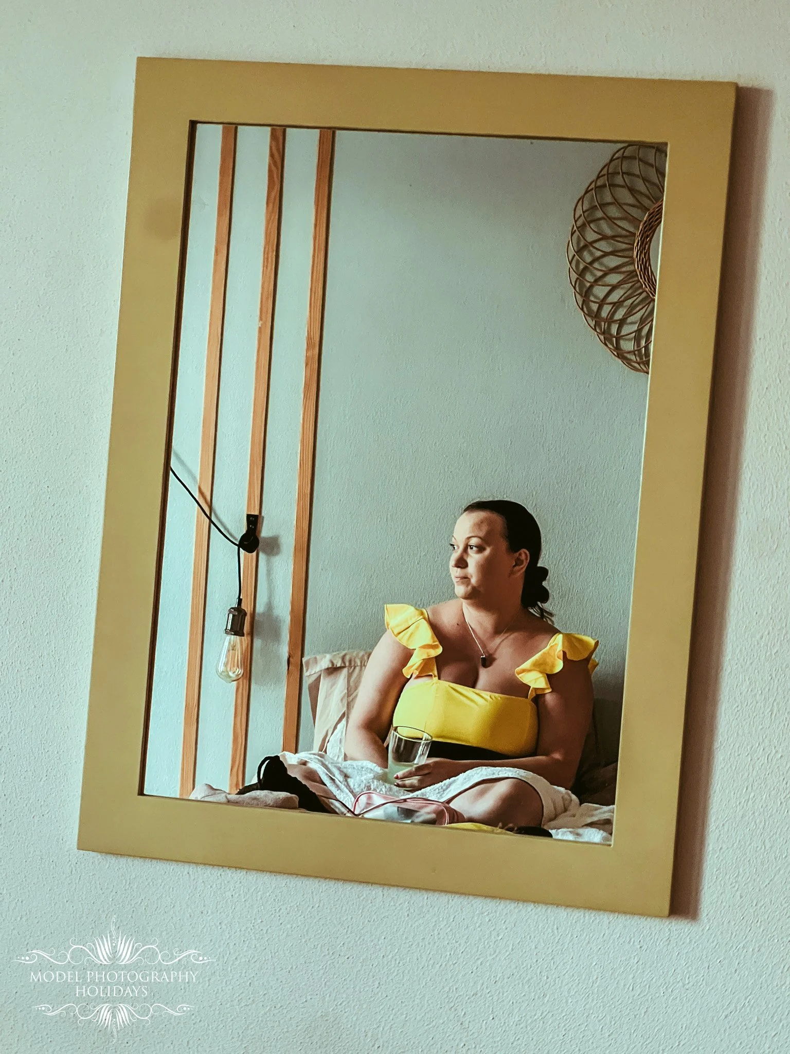 A woman in a yellow top is sitting on a bed holding a glass, reflected in a mirror on the wall.