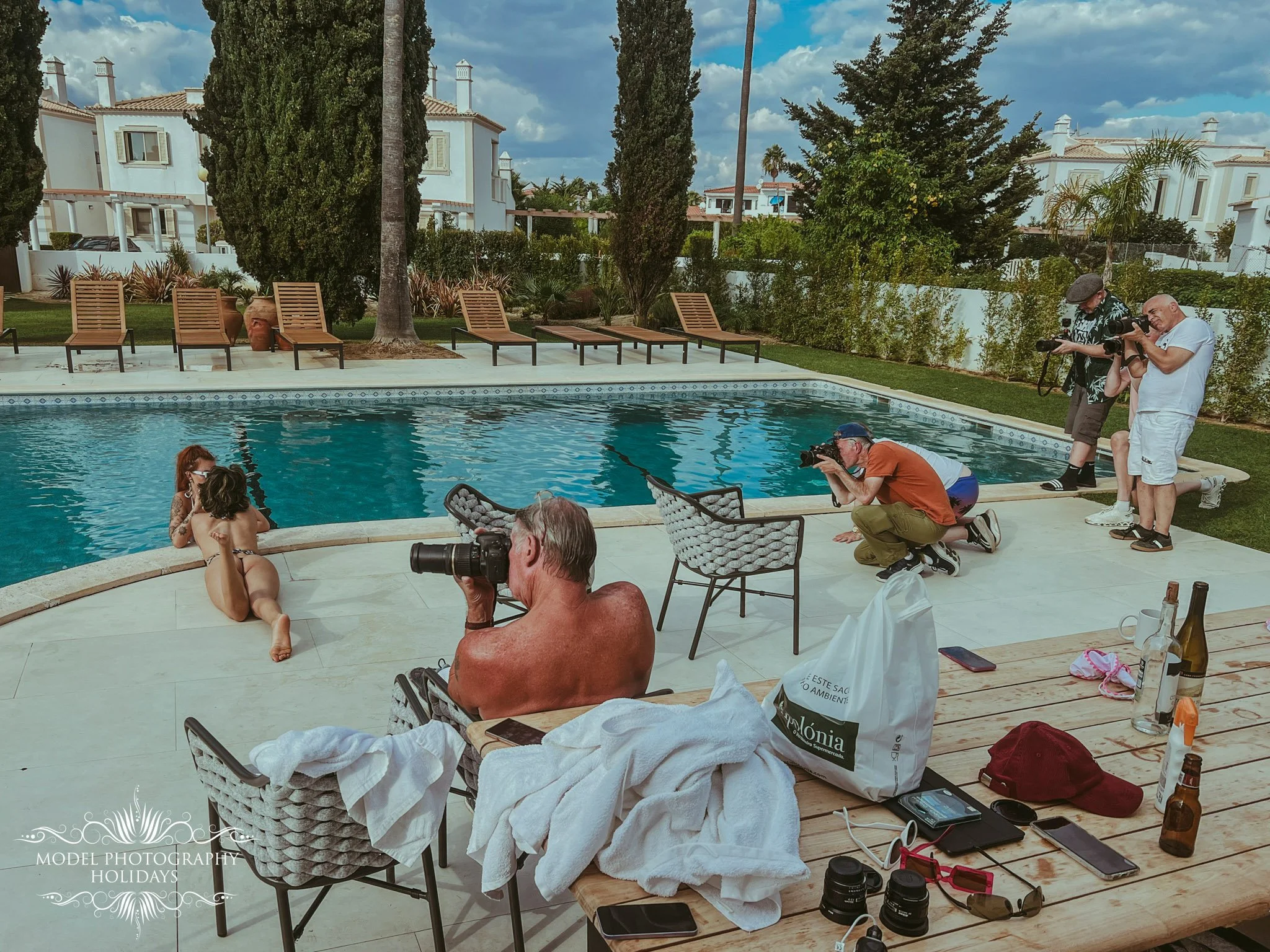 People participating in a photo shoot by a swimming pool at a house with trees, lounge chairs, and a partly cloudy sky in the background.