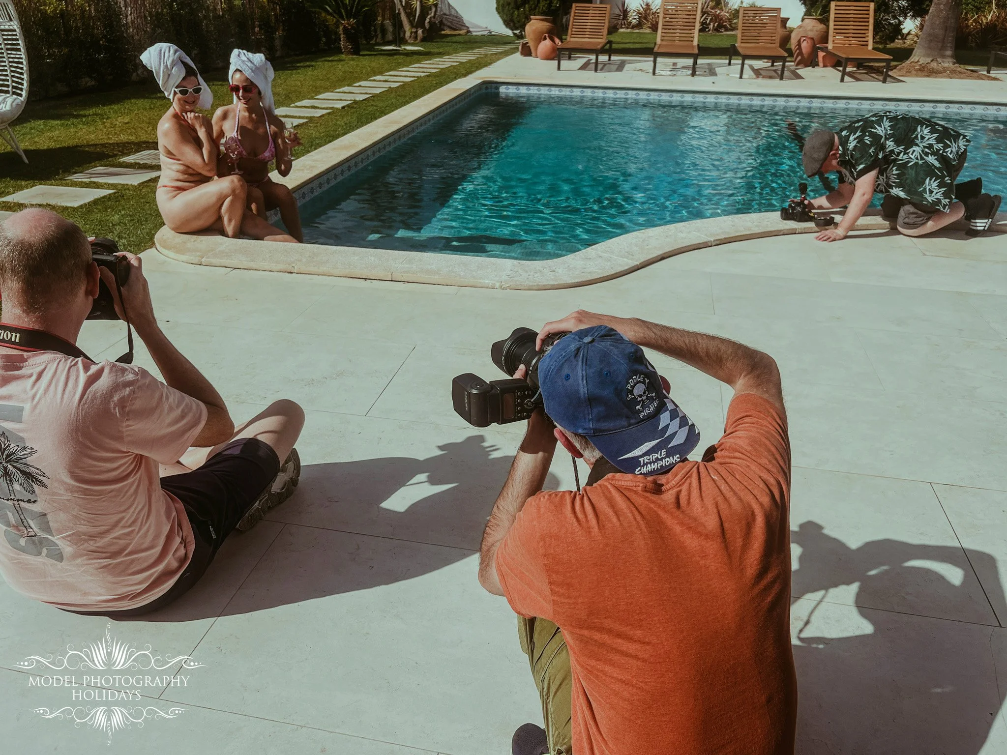 Two women sitting by a swimming pool with towel turban on their heads, smiling and wearing sunglasses, photographed by two men holding professional cameras. In the background, there are lounge chairs and potted plants. The scene depicts a relaxing ou
