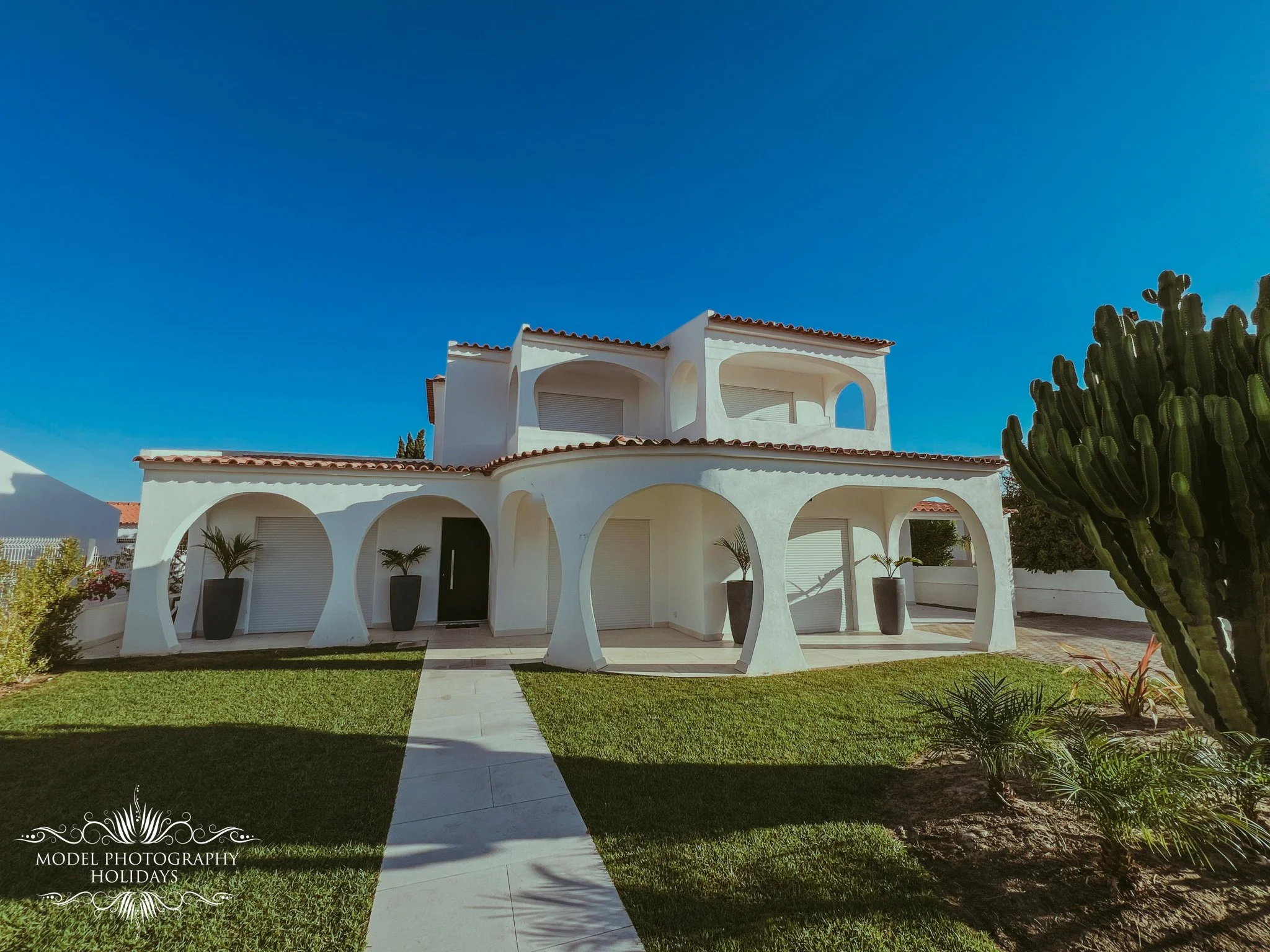 White modern house with arched balconies and terracotta roof tiles, surrounded by green lawn and desert plants, under a clear blue sky.