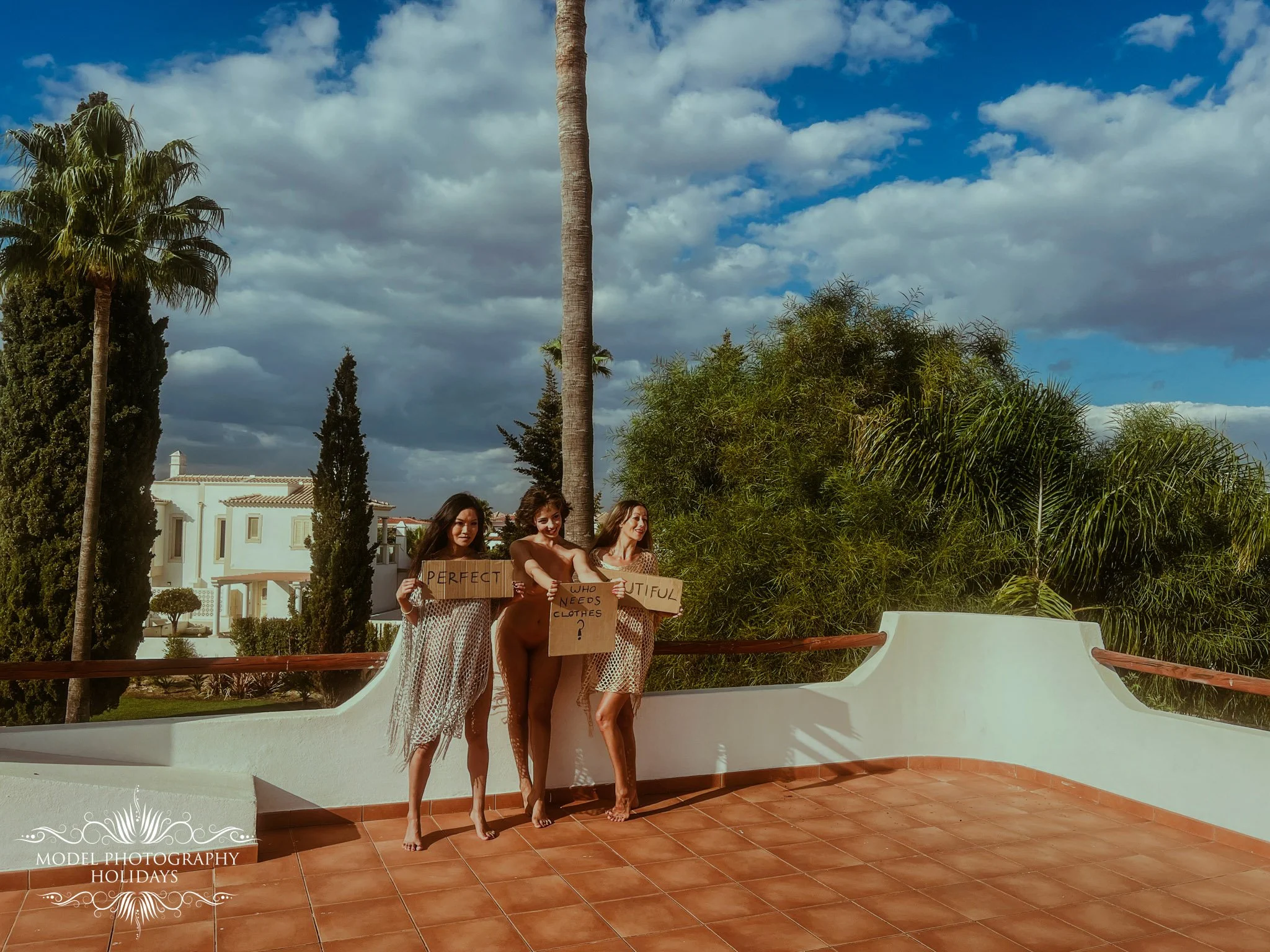Four women in swimsuits and cover-ups are standing on a balcony with a white wall, holding cardboard signs with messages. They are smiling and looking at each other or the camera. Behind them are palm trees, lush greenery, and a cloudy sky. The image