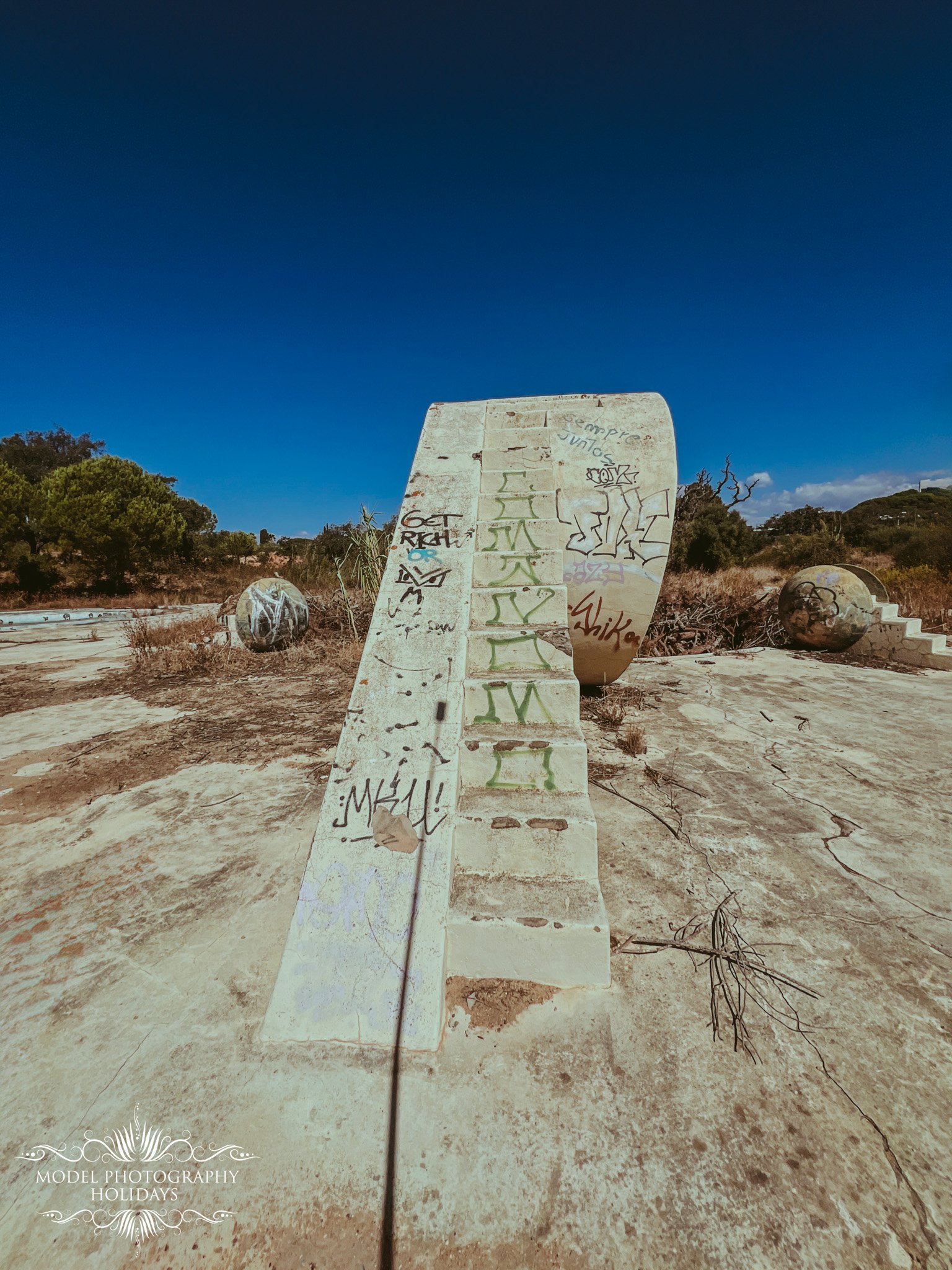 A large, weathered, white concrete sculpture with graffiti and writings on it, surrounded by smaller spherical sculptures with graffiti, in a dry, open area with a clear blue sky and green trees in the background.