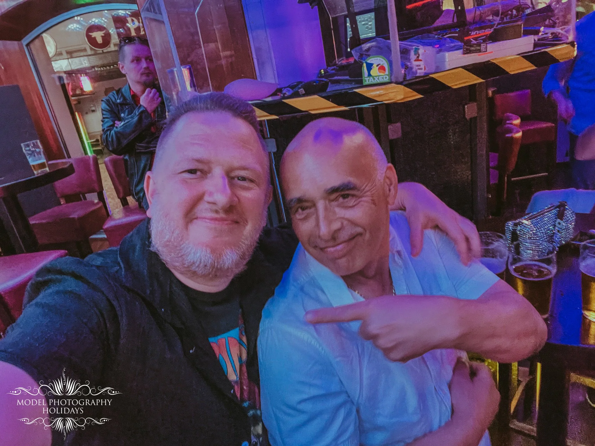 Two men taking a selfie in a colorful bar or club with purple and pink lighting. One man has a beard and is wearing a dark shirt; the other man is bald and wearing a light-colored shirt. There are tables, chairs, and a person in the background near a