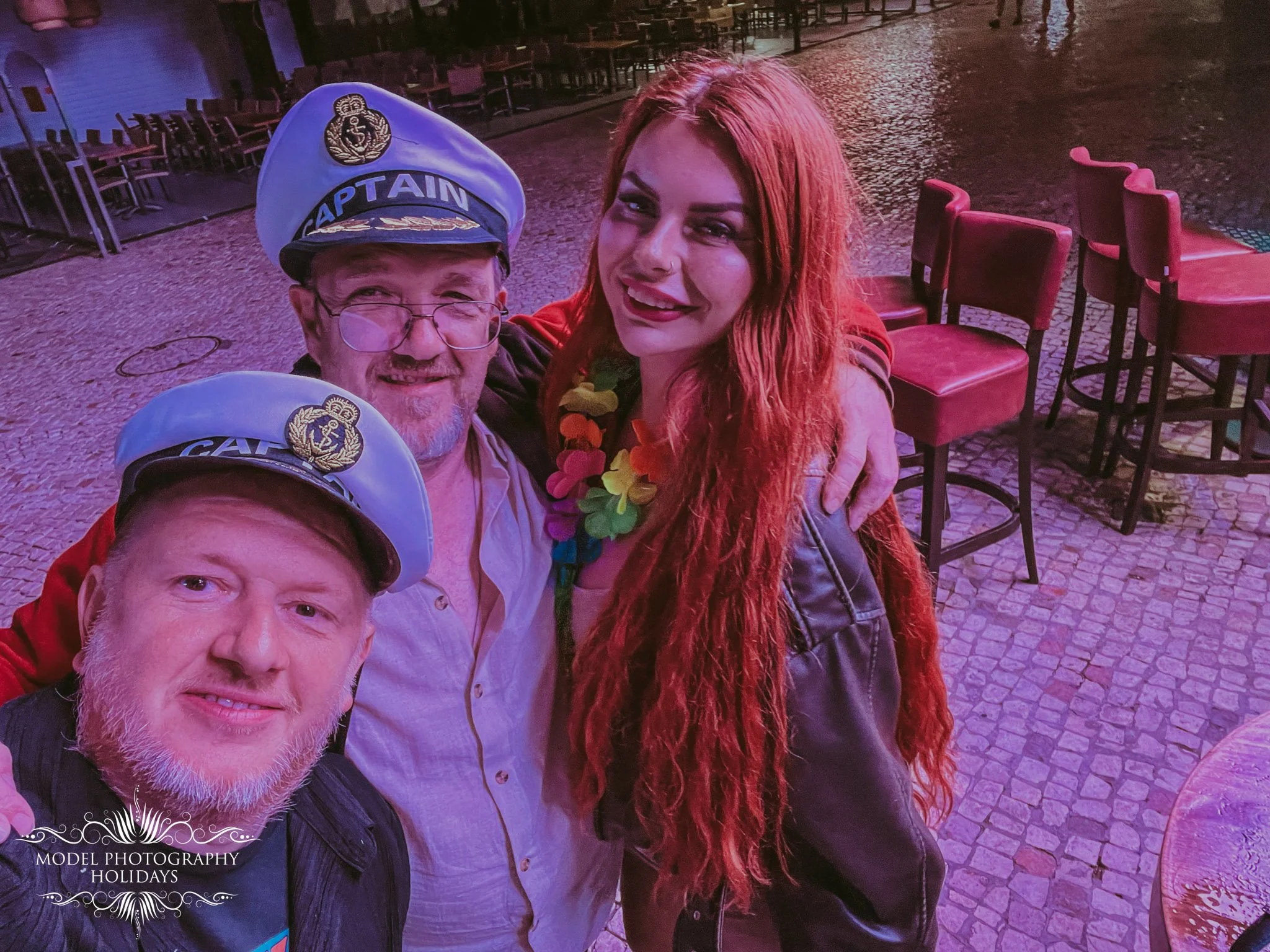 Group of four people taking a selfie on a cobblestone street at night, with empty chairs and tables in the background, and logo for Model Photography Holidays in the bottom left corner.
