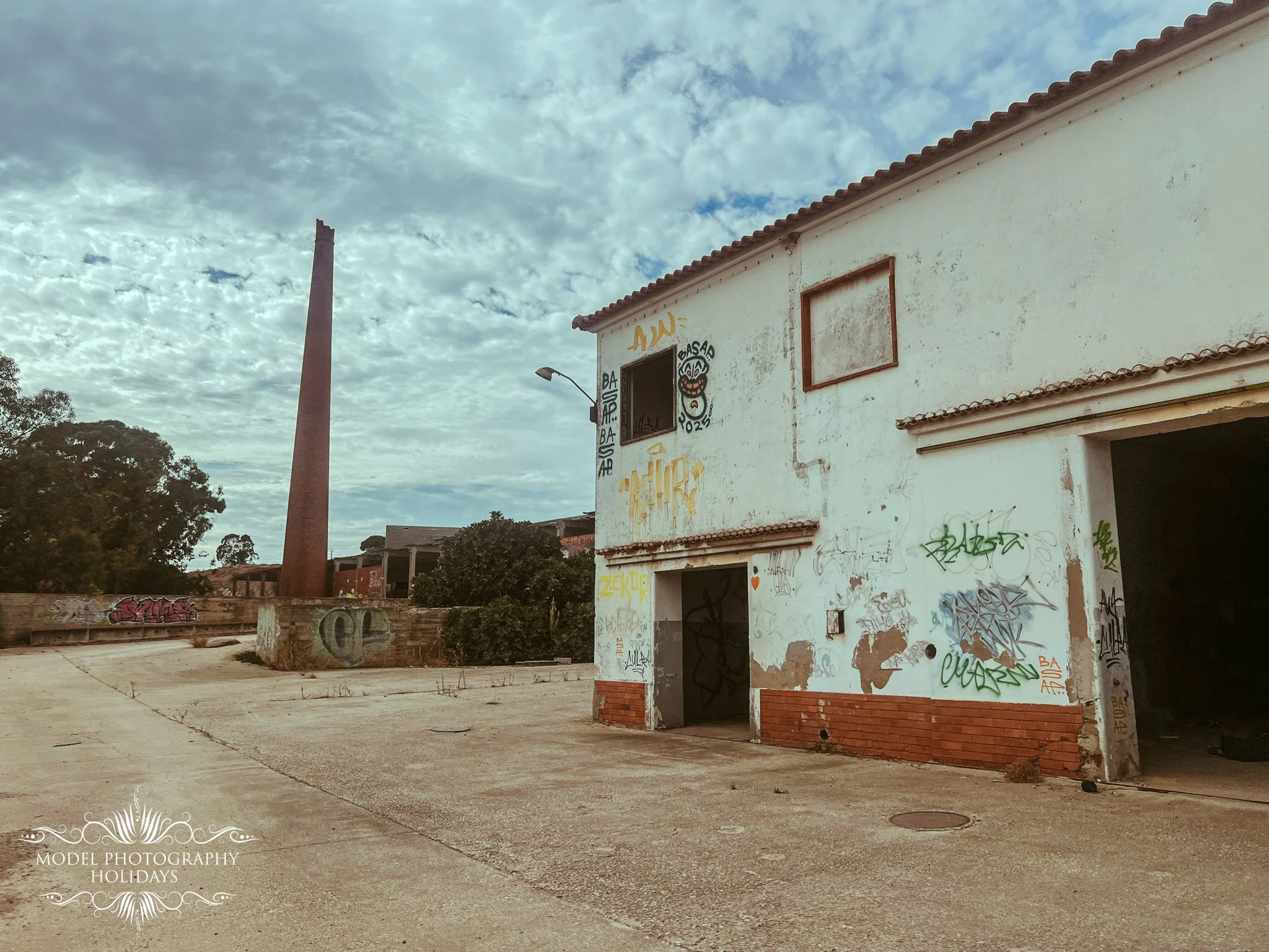 An empty, partially boarded-up building with graffiti on the walls, located in an open area with a chimney to the left against a cloudy sky.