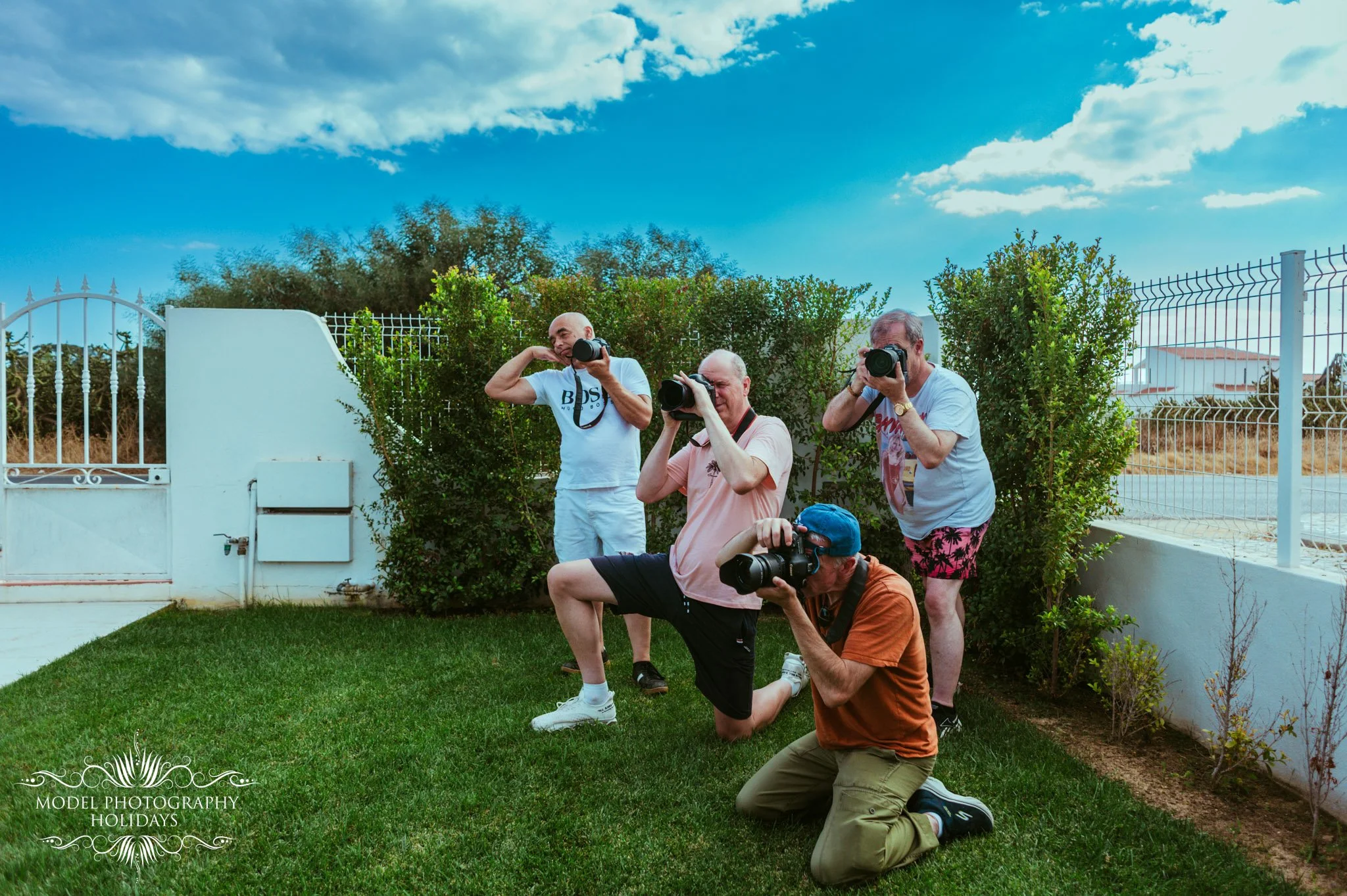 Four men taking photographs outdoors in a garden with green bushes and a blue sky with clouds. The men are in different poses, using professional cameras, and are dressed casually.