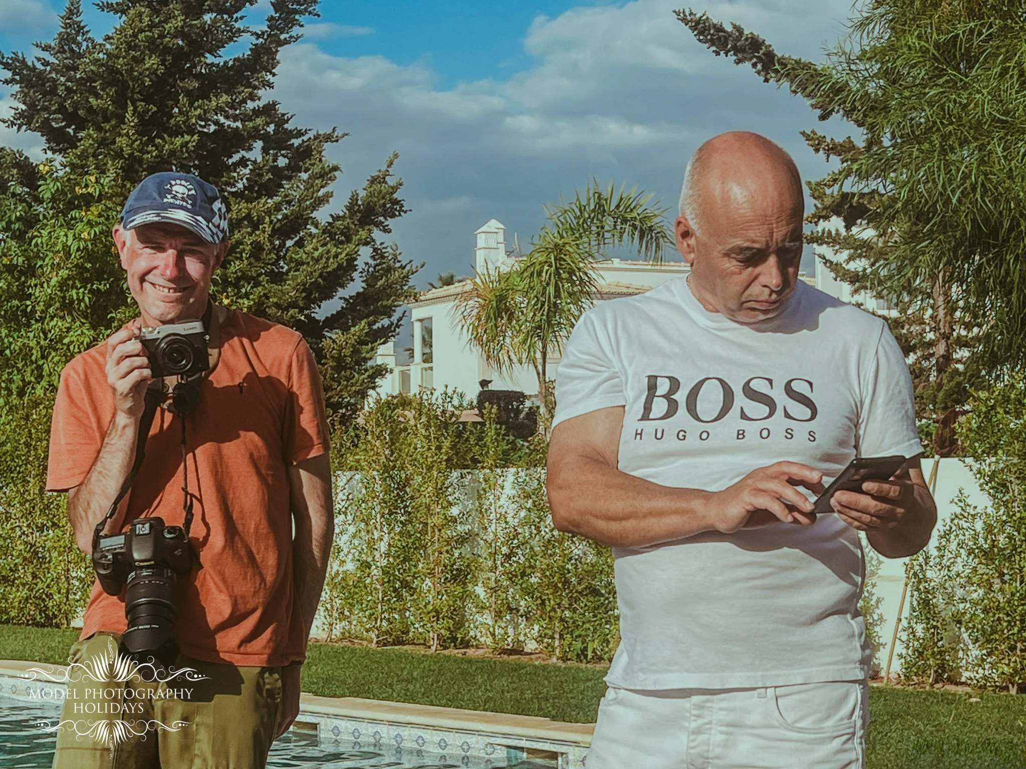 Two men standing outdoors among trees and shrubs, one smiling and holding a camera, the other looking at a smartphone, with a building and cloudy sky in the background.