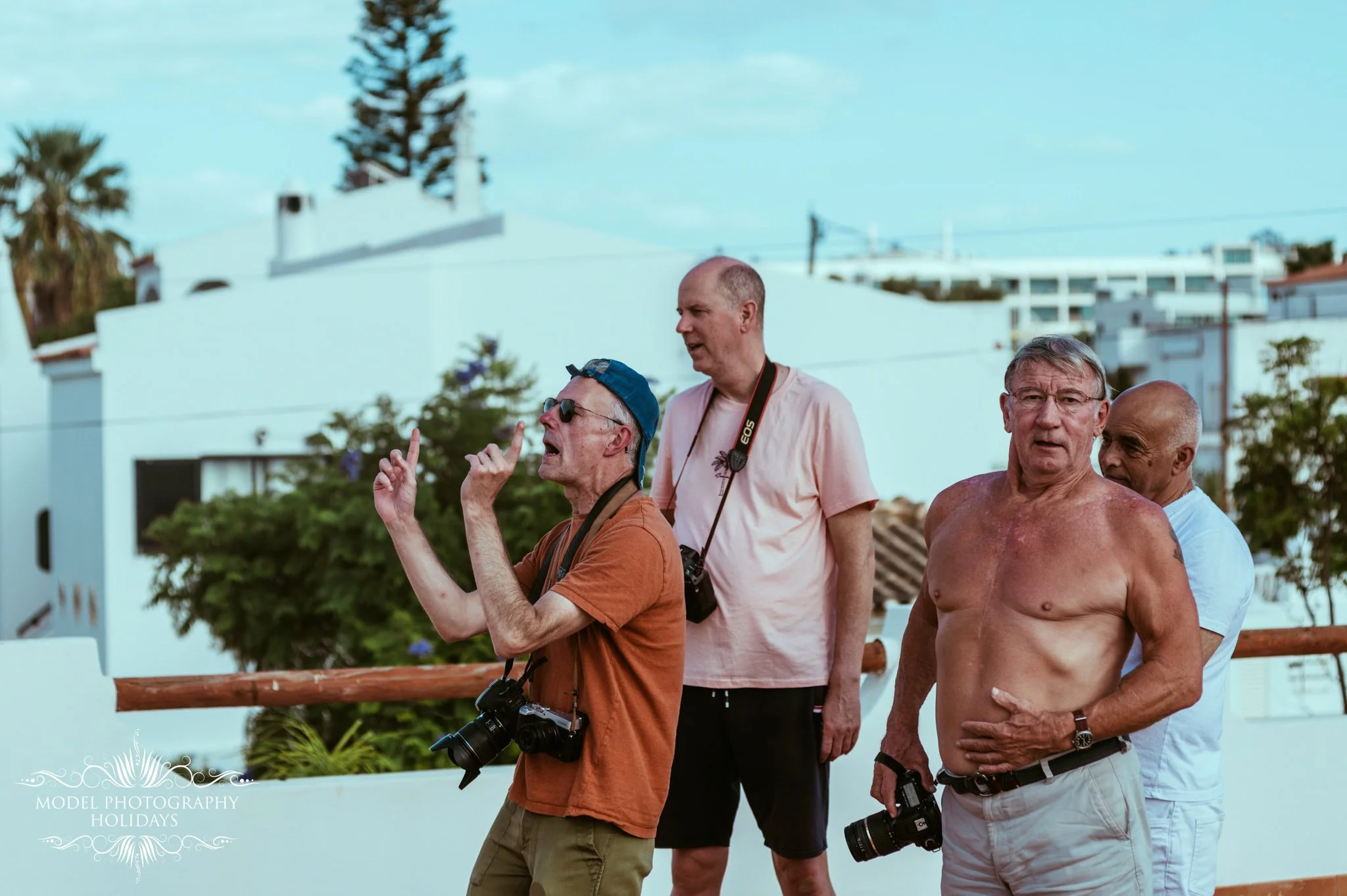 Four men are walking outdoors in a sunny area with white buildings and a blue sky in the background. One shirtless older man is among the group, and two of them have cameras around their necks.