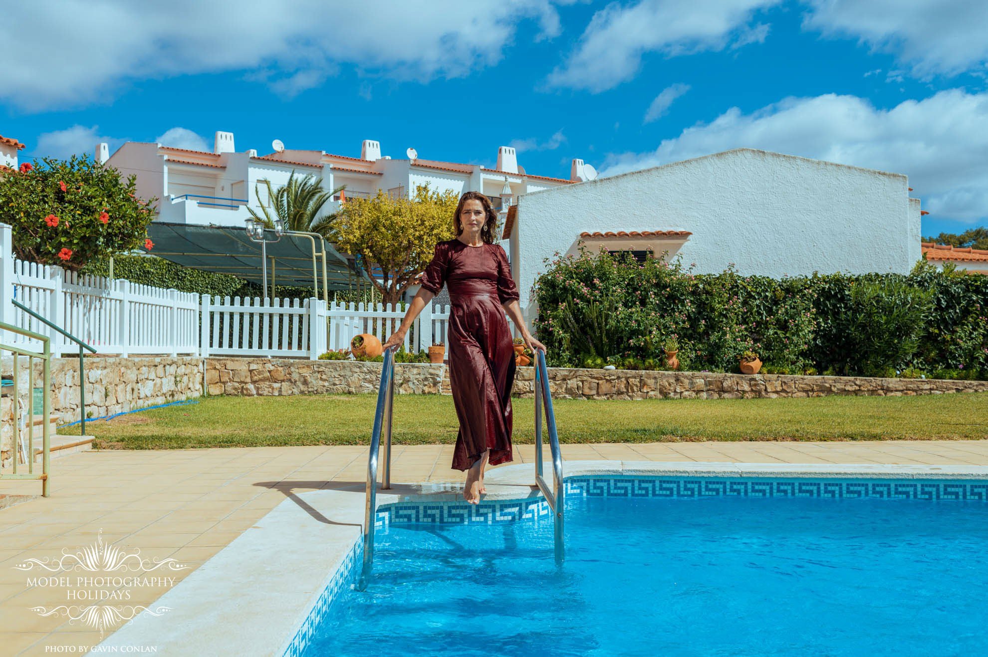 Woman in red dress descending pool steps in a sunny garden with white houses in the background.