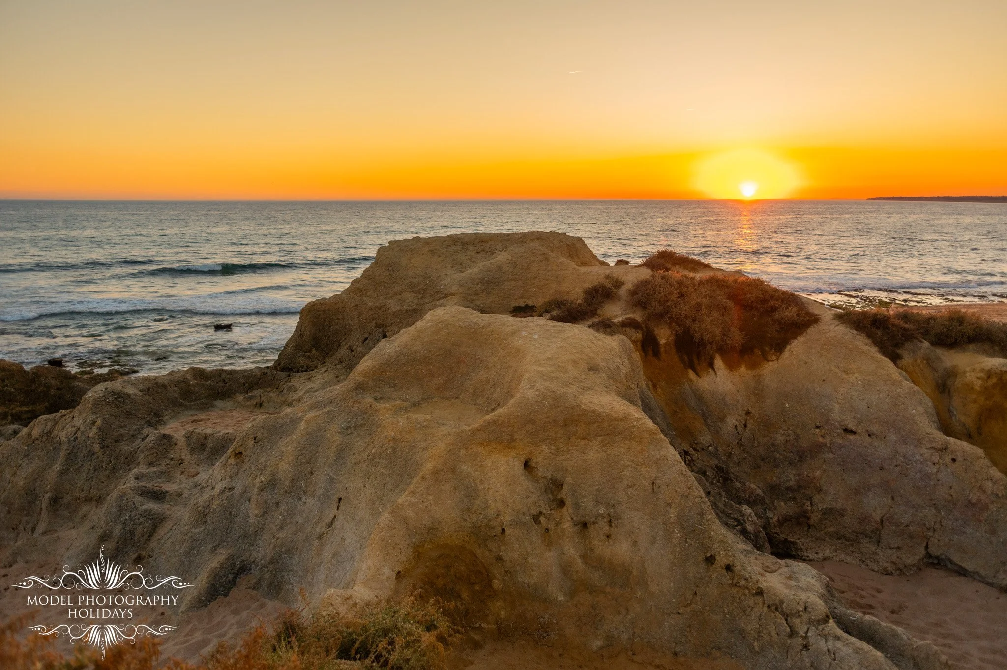 Sunset over the ocean with rocky coastline in the foreground.
