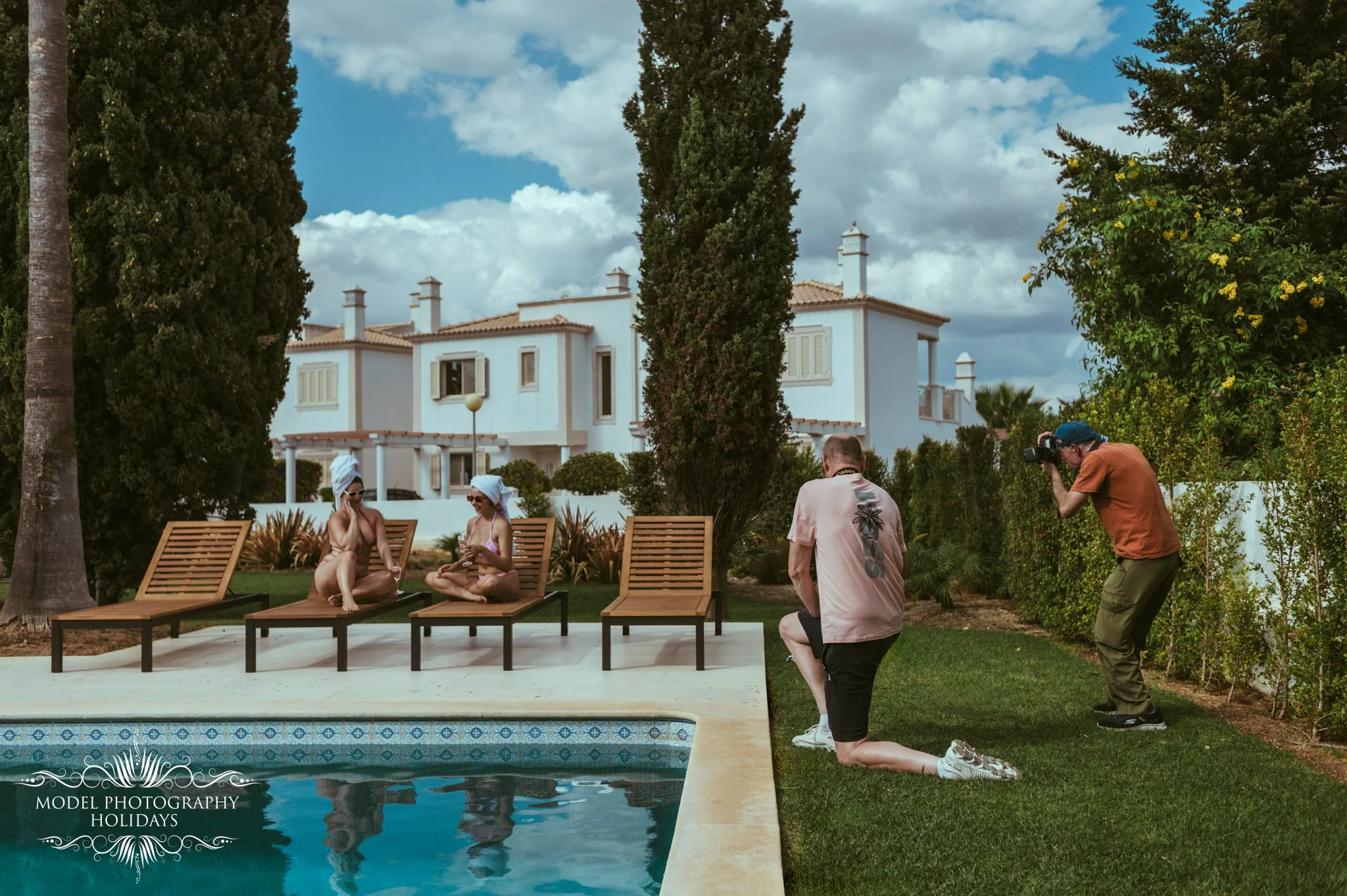 Photographers taking pictures of two women sitting by a swimming pool at a sunny estate with a white house and tall trees in the background.