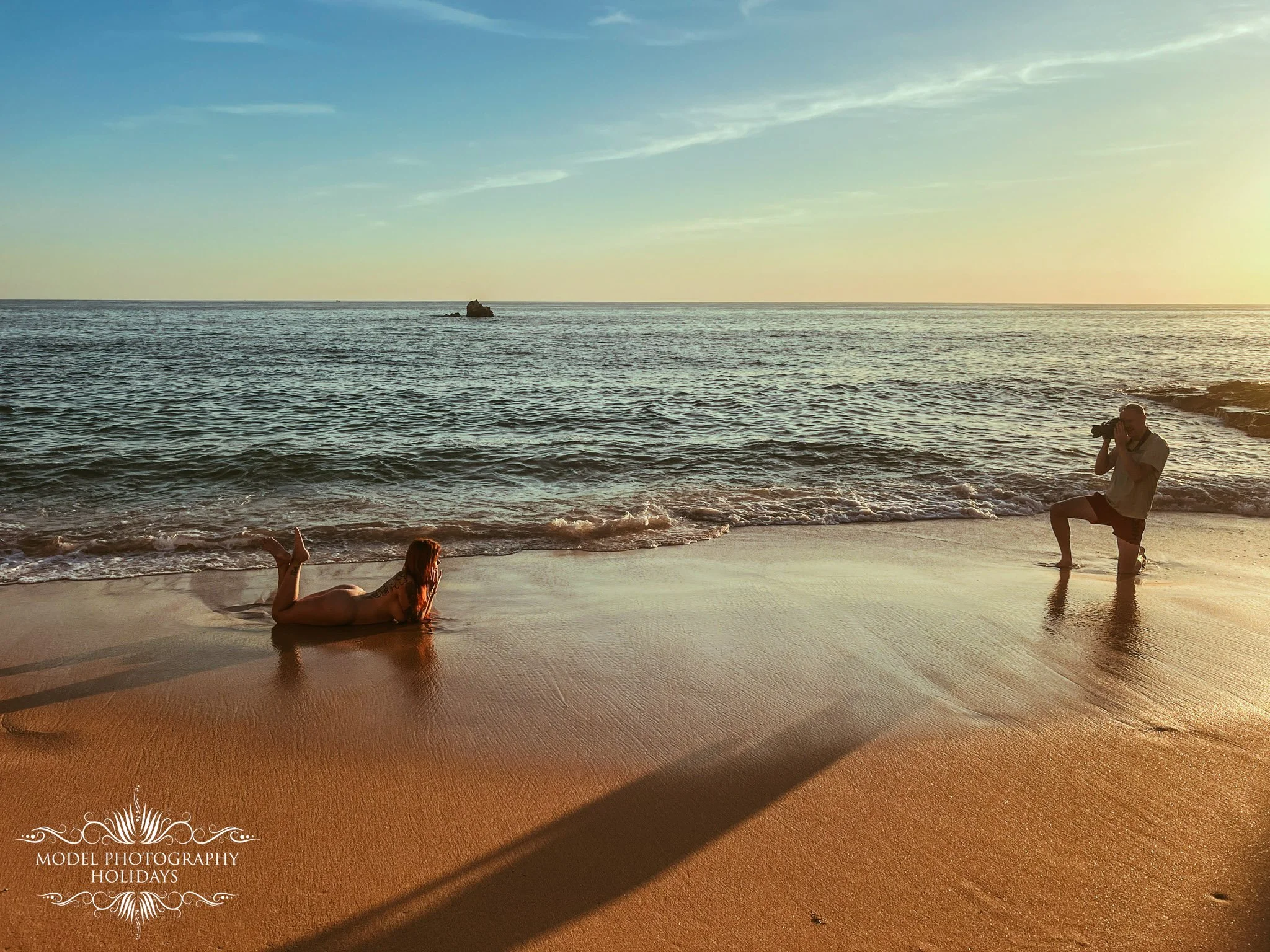 A person lying on the beach with a sunset in the background, another person kneeling by the water taking a photo, and a photographer in the right foreground.