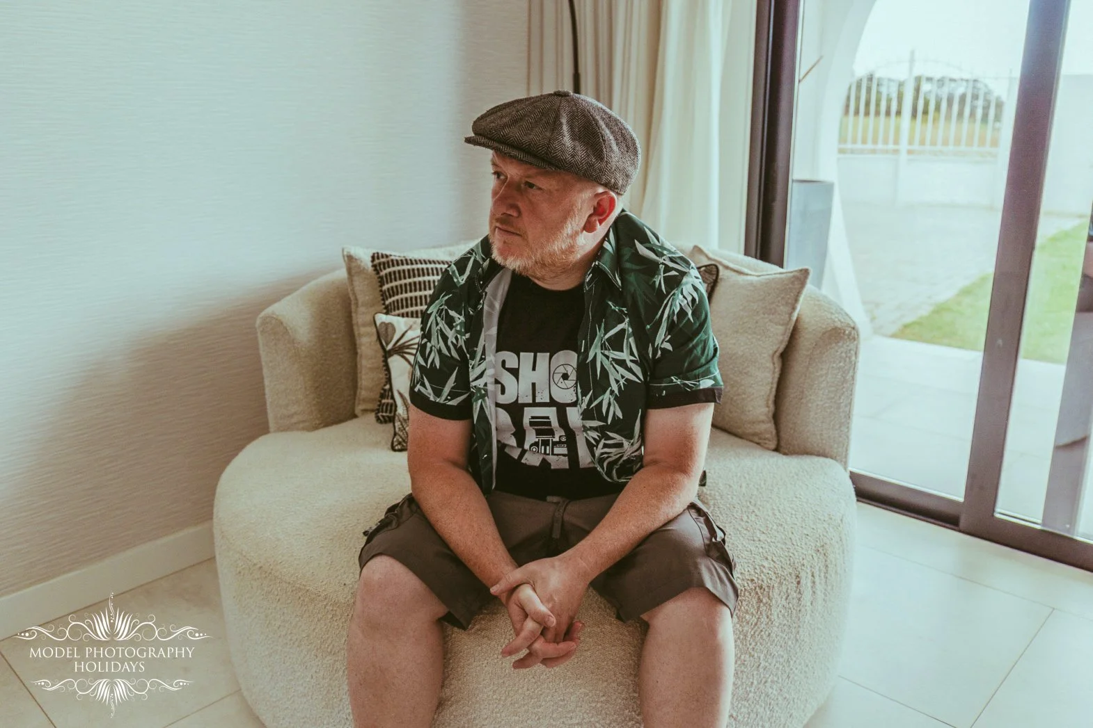 A man sitting on a beige couch in a sunny room, wearing a gray flat cap, a graphic T-shirt, and shorts, looking to his left near a sliding glass door with a view of a yard.