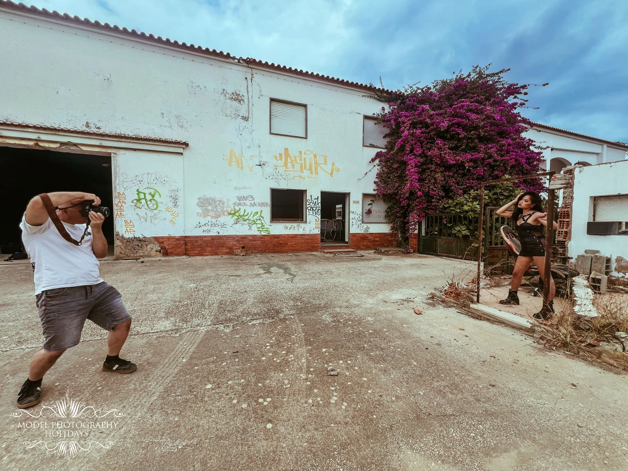 A man with a camera taking a photo in an outdoor setting with a woman posing near a large purple flowering bush and an old white building with graffiti in the background.
