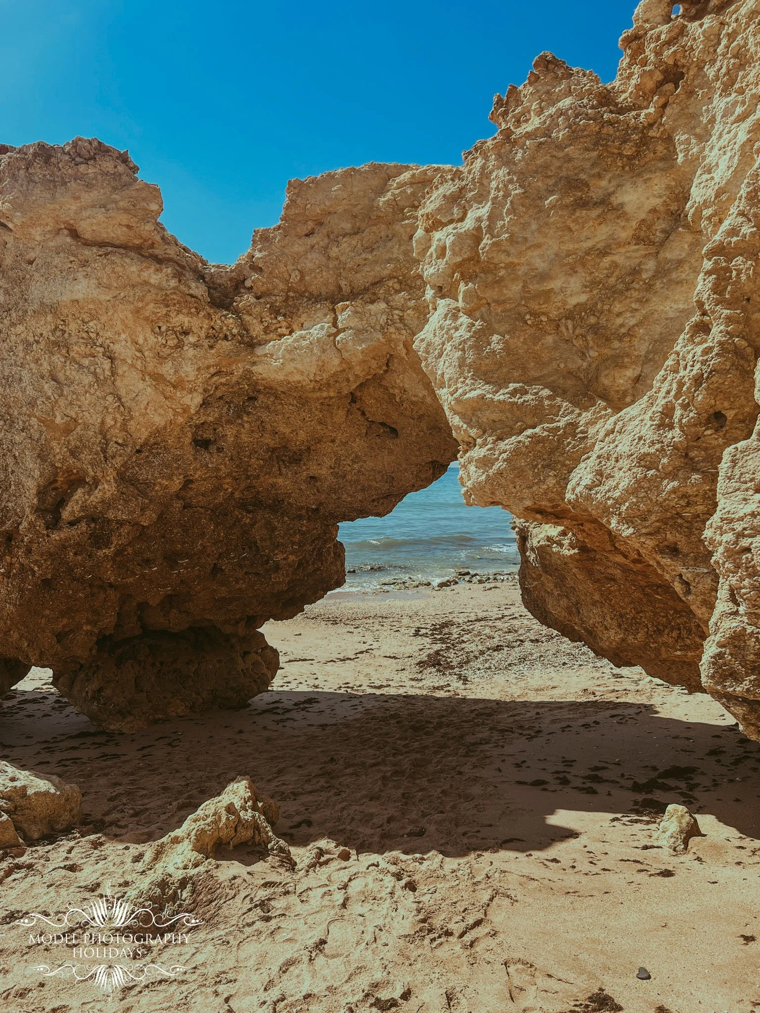 View through a natural rock formation to sandy beach and blue ocean water with a clear sky.