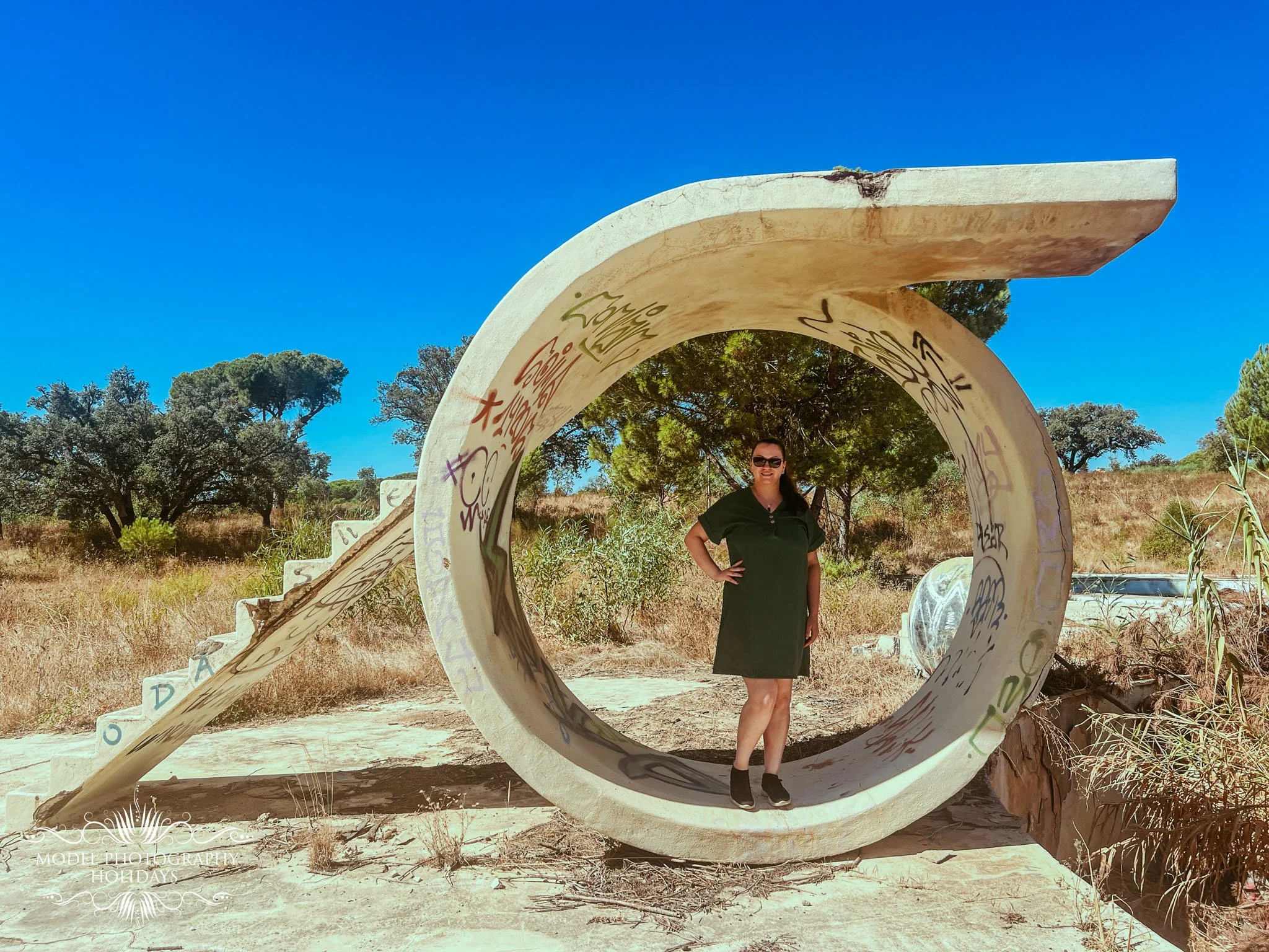 A person standing under an artistic concrete arch with graffiti in a natural outdoor setting, trees and blue sky in the background.