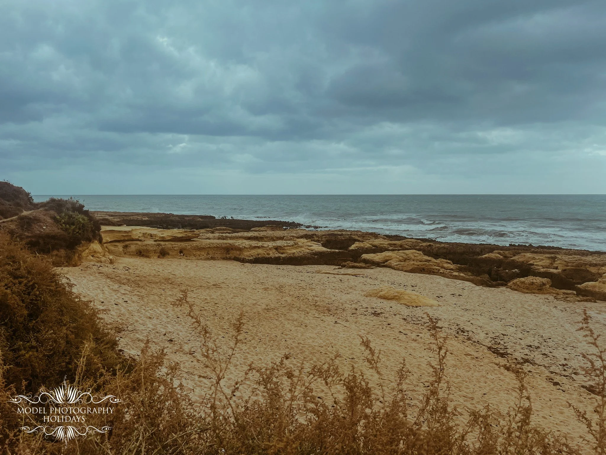 A sandy beach with some rocks and sparse vegetation, overlooking the ocean with cloudy skies.