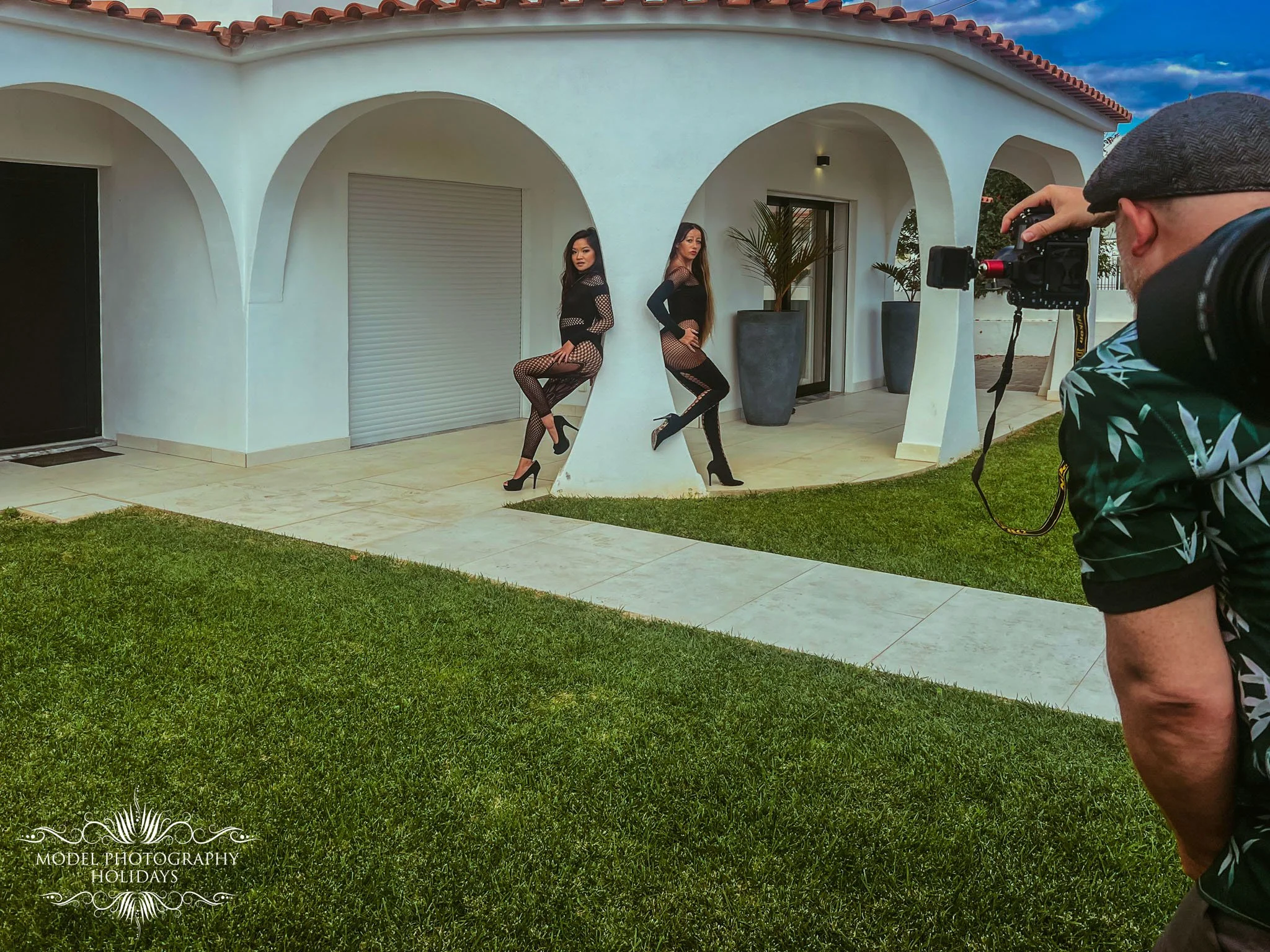 Two women posing in black fishnet outfits with high heels, standing against a white building with arches, next to large potted plants. A man is taking their photograph with a camera, wearing a cap and patterned shirt.