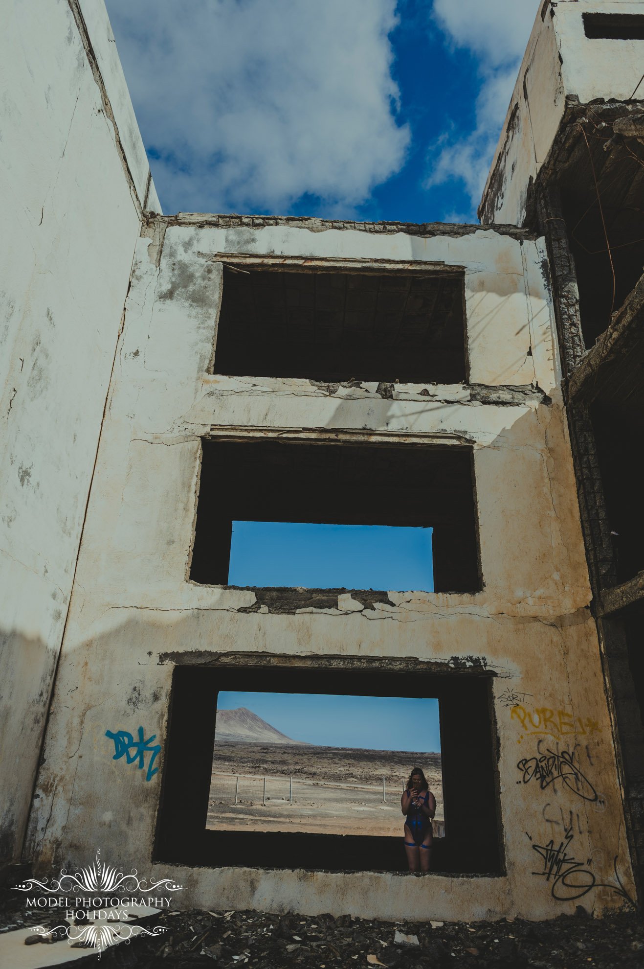 Abandoned building with large windows, graffiti on walls, blue sky visible through openings, person standing in lower window overlooking desert landscape.