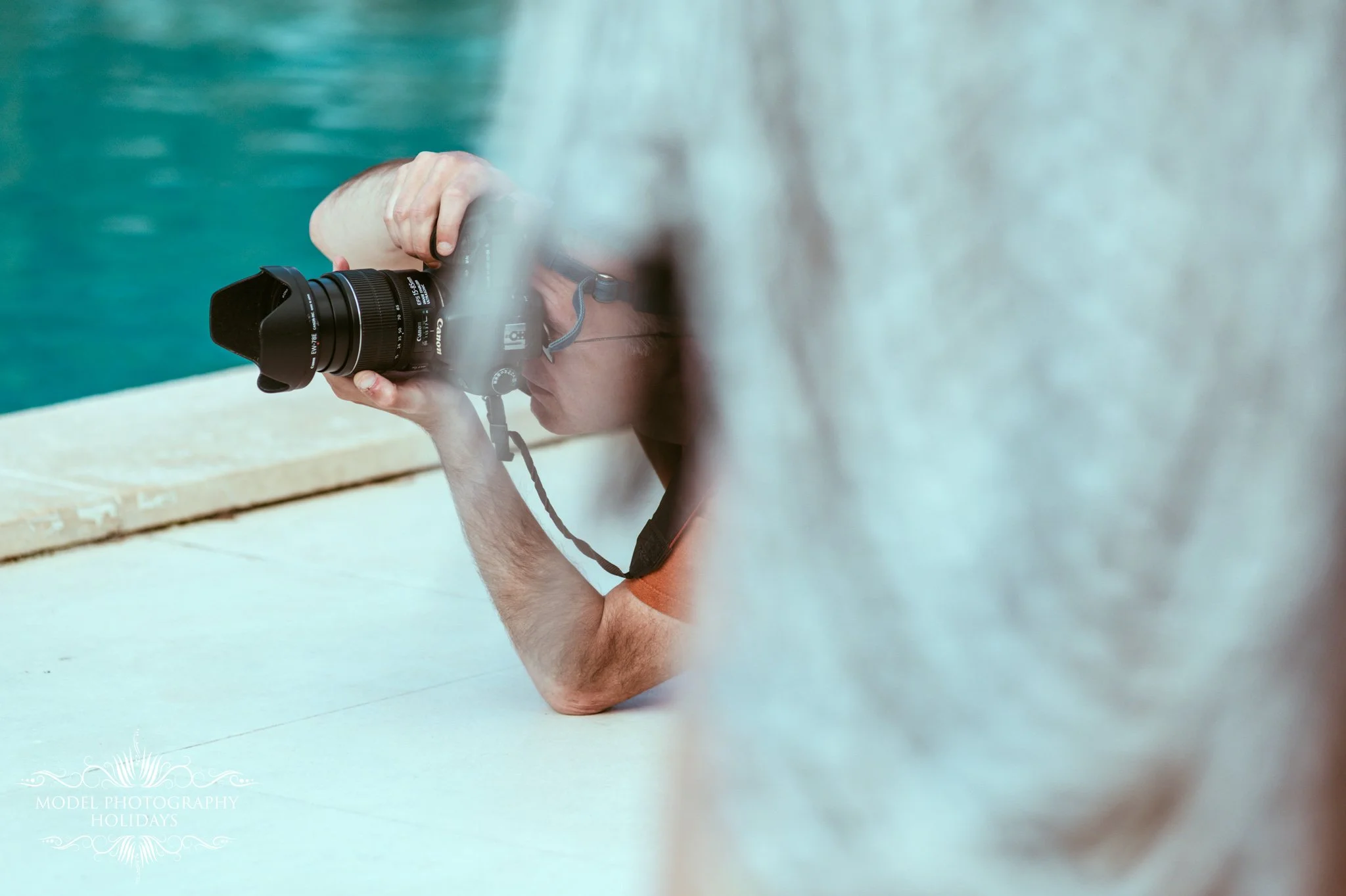 A person taking a photograph with a Canon camera while lying on the ground, with a blurred foreground and a blue water body in the background.