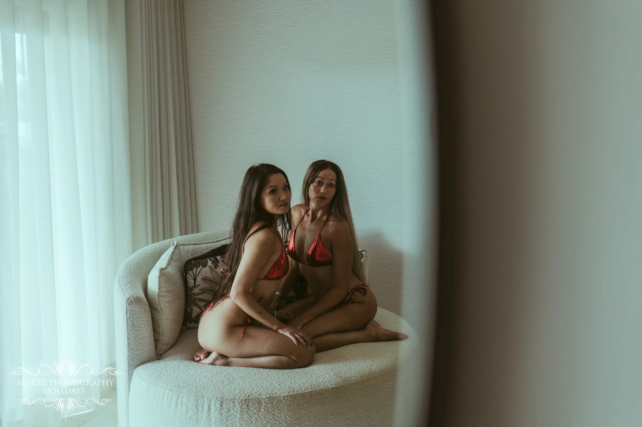 Two women sitting on a beige couch in a room with off-white walls and vertical blinds. Both women are wearing red satin lingerie and sitting closely, with serious expressions.