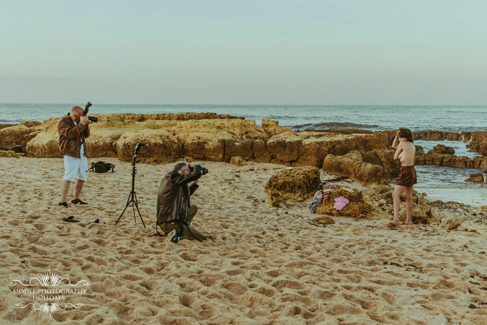 A photoshoot at the beach with a young woman in a bikini, two photographers taking pictures of her, and the ocean in the background.