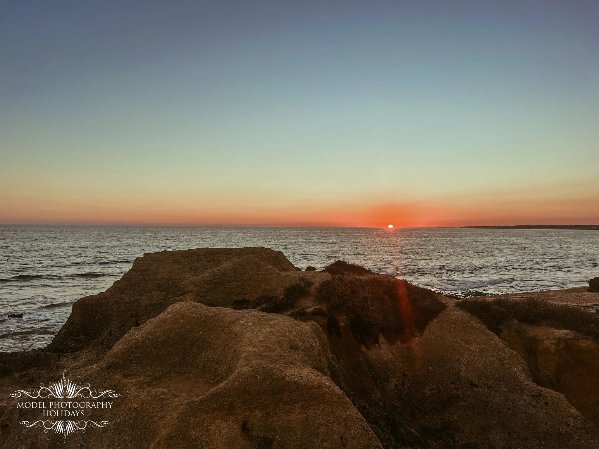 Sunset over the ocean with rocks in the foreground and a clear sky.
