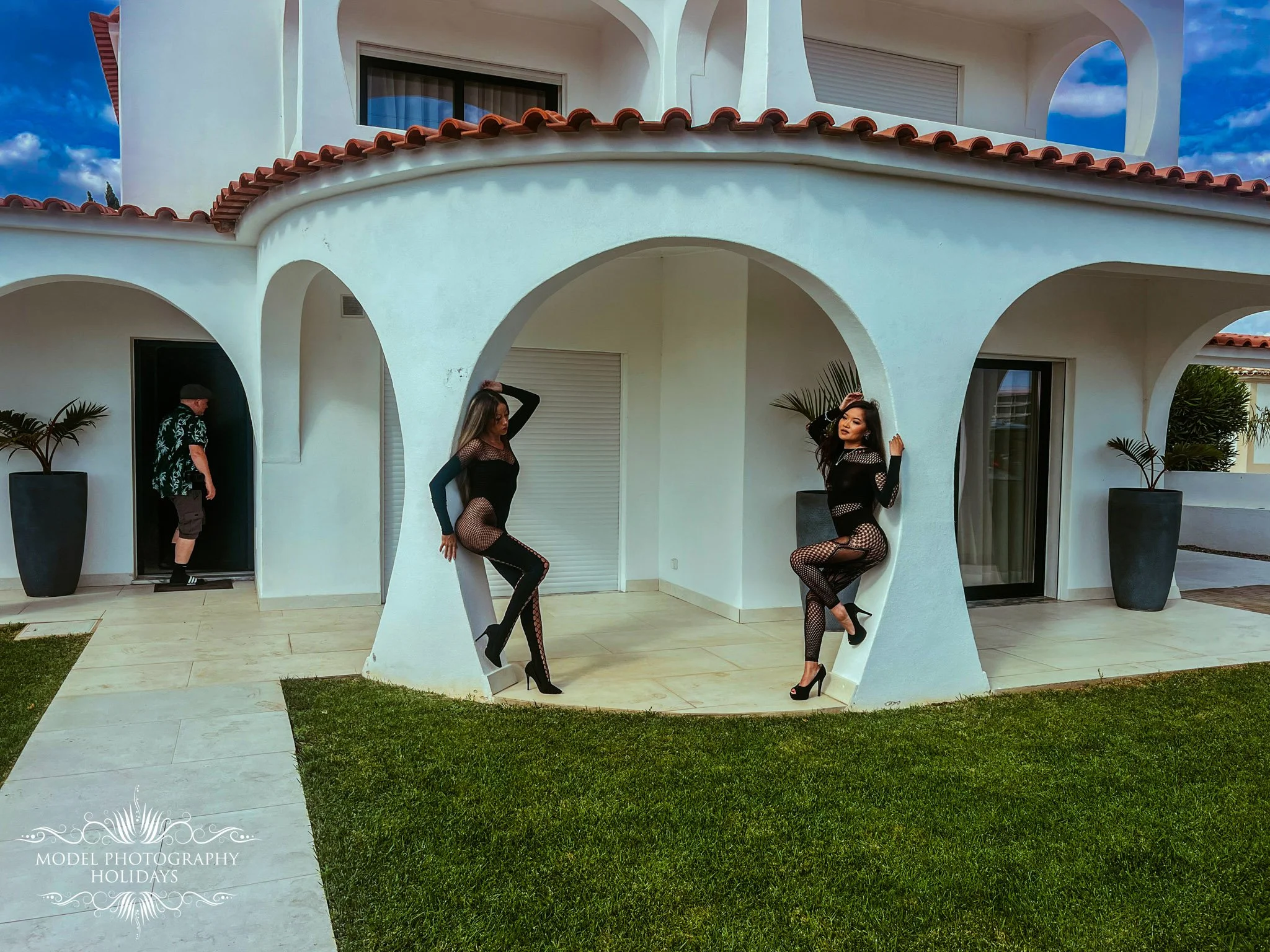 Two women in black fishnet dresses and high heels posing near a white modern architectural structure featuring arches and curved walls, with a man in the background near the door of a white house with a tiled roof.