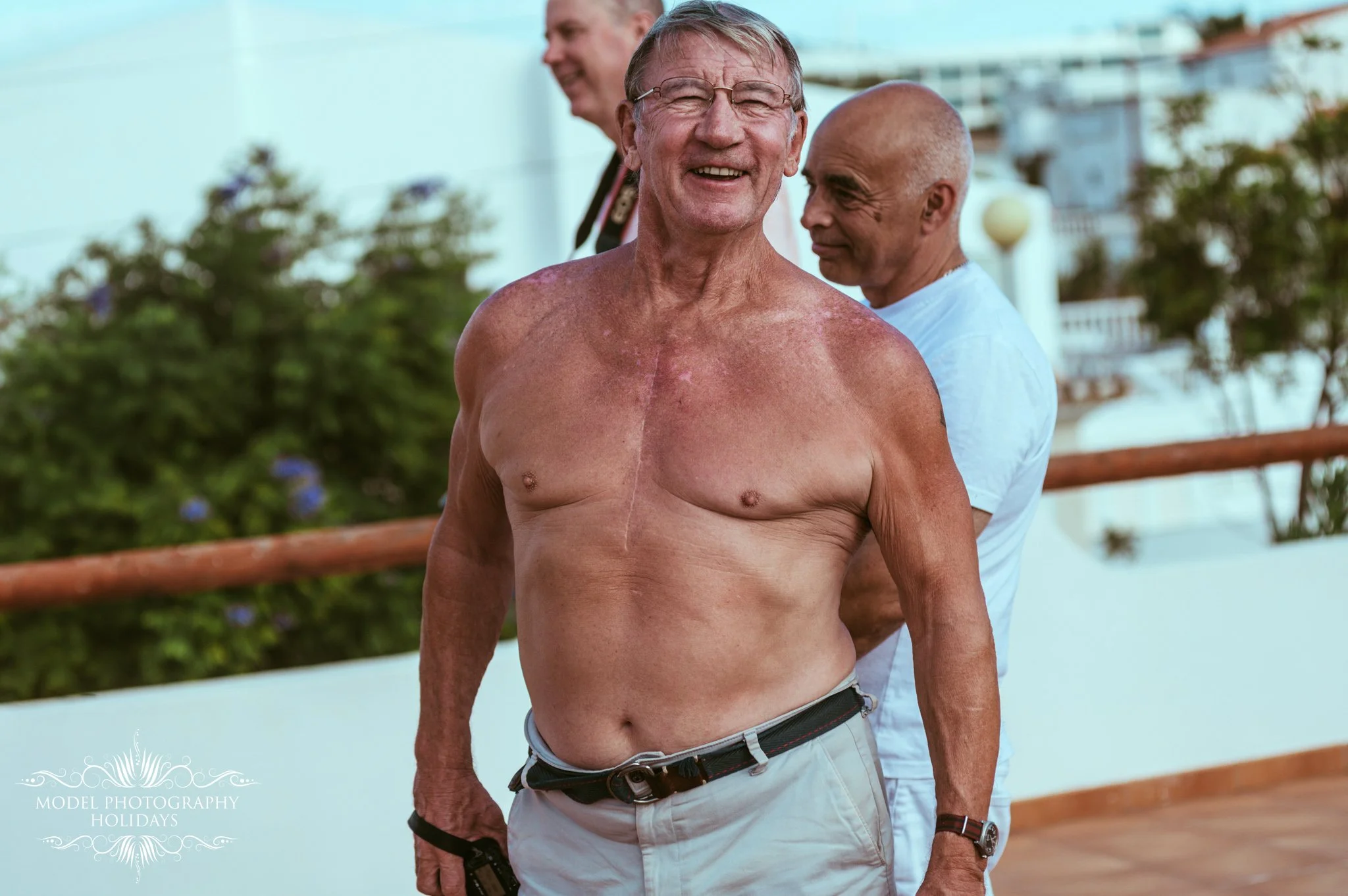Smiling elderly man with glasses and tan skin, shirtless, standing outdoors on a wooden deck with a woman behind him and another man in the background, surrounded by greenery and a white building.
