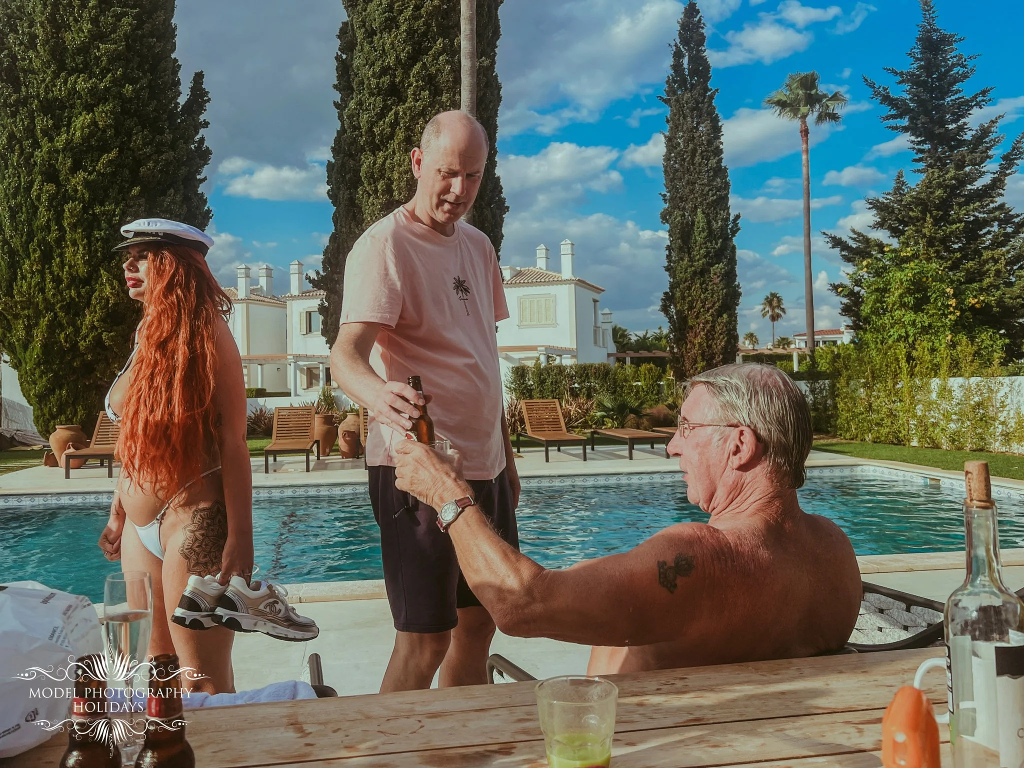 People at a poolside gathering with trees and houses in the background, some standing, some seated, with drinks and food on the table.