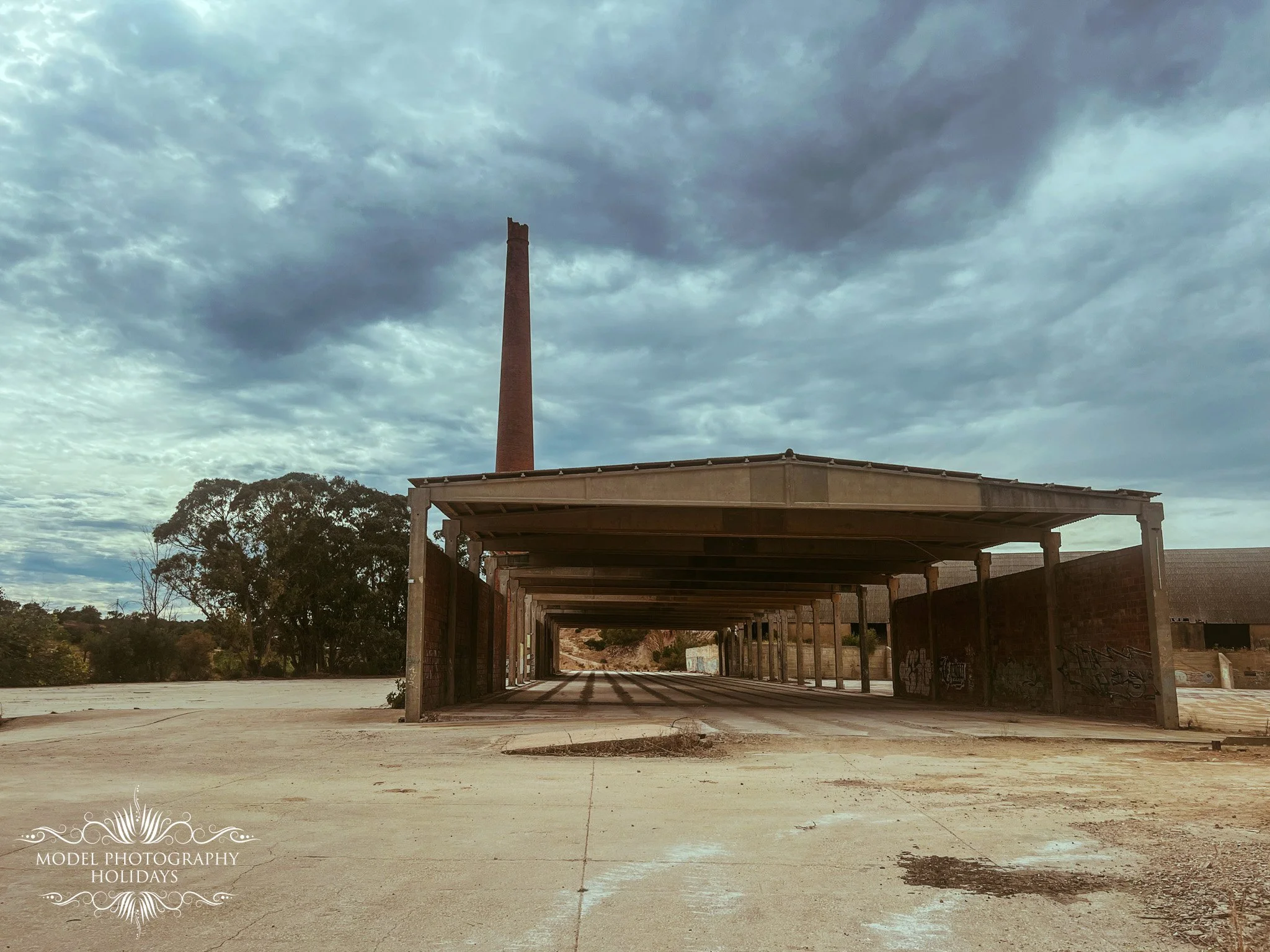 An outdoor scene featuring an under-construction building with a roof structure supported by wooden beams. A tall, narrow brick chimney rises into a cloudy sky. There are trees on the left side and some graffiti on a wall in the background.