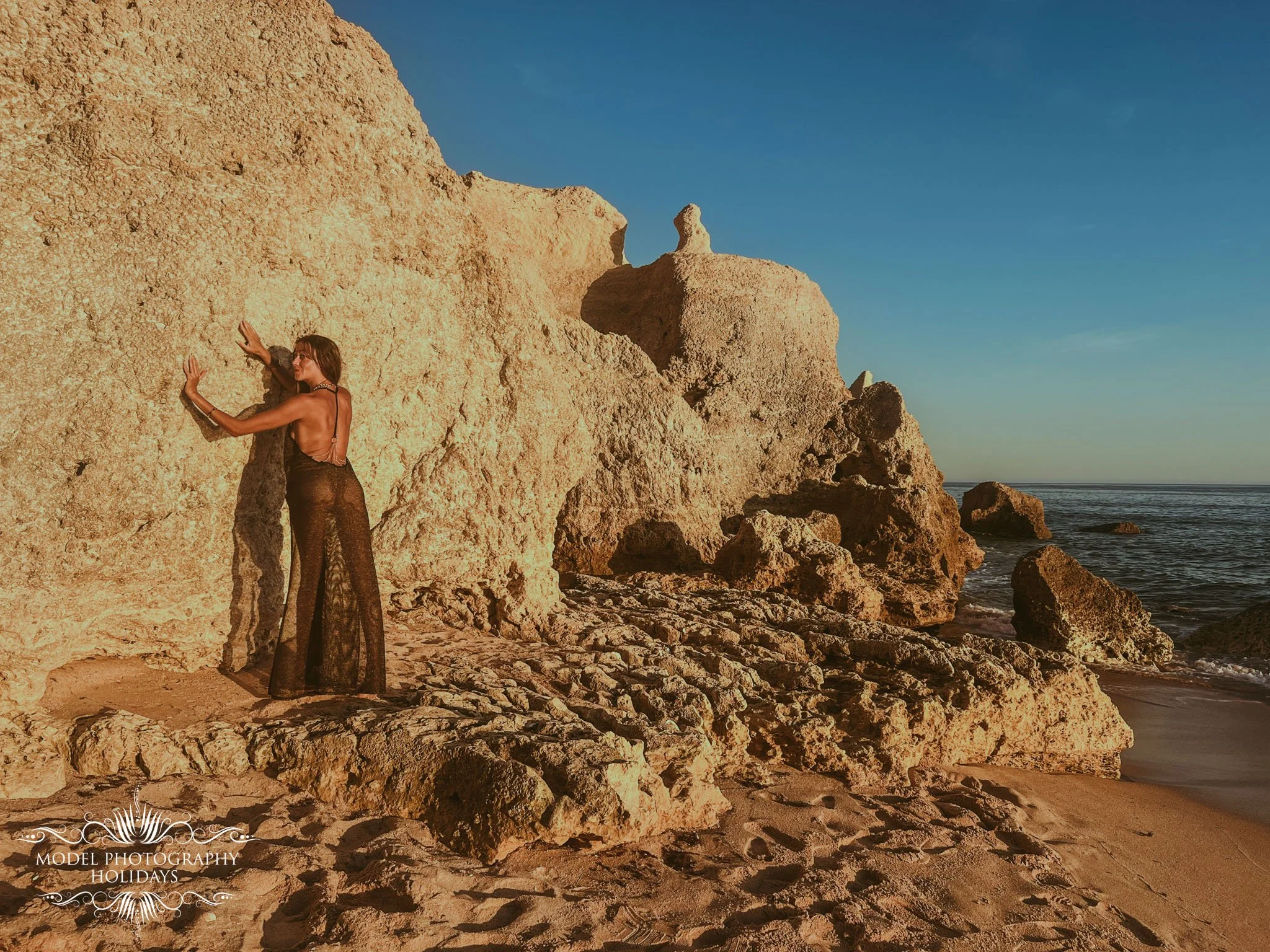 Woman in a sheer black dress poses against large rocky formations on a beach at sunset, with the ocean in the background