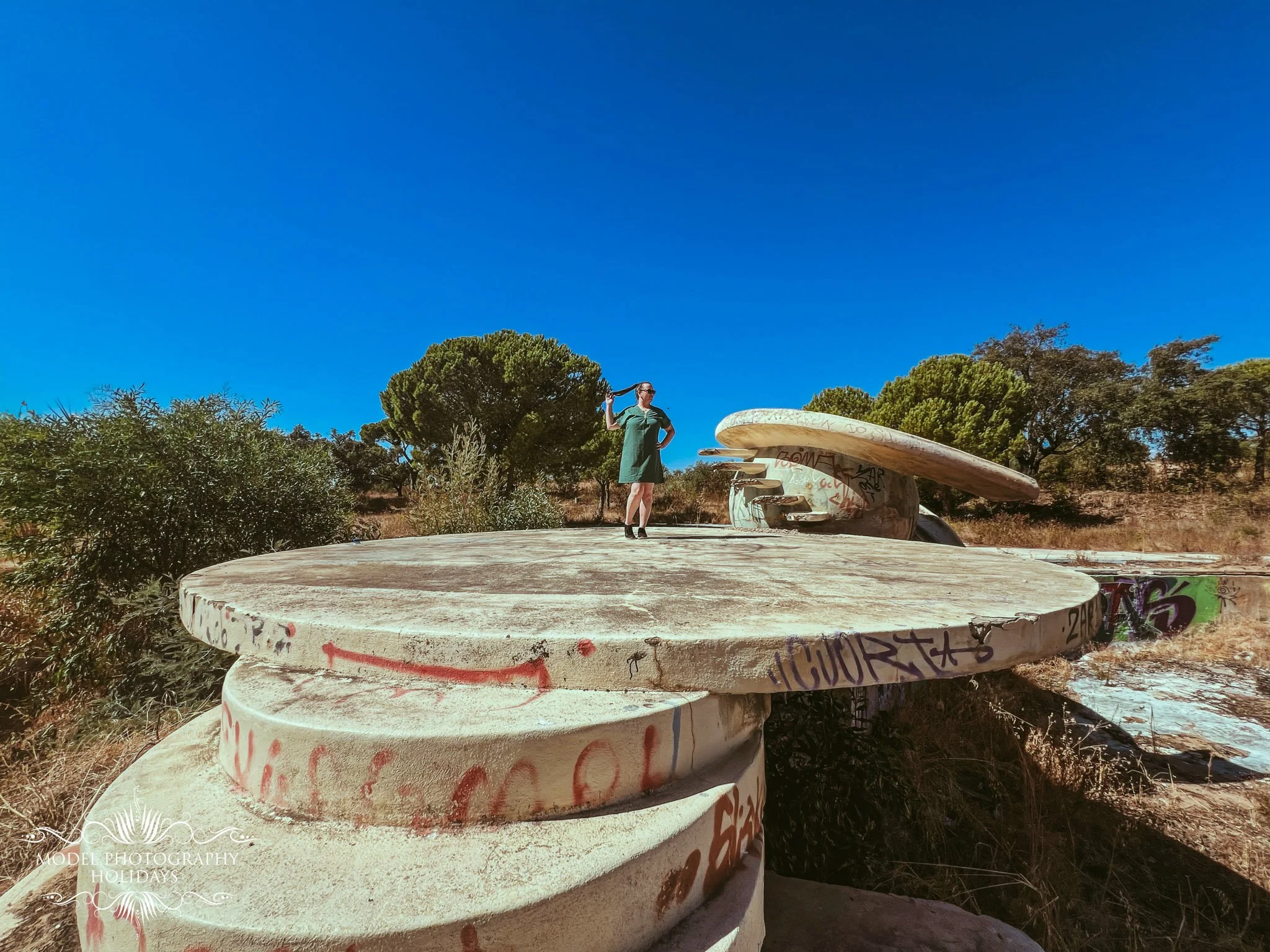 A woman in a green dress and sunglasses standing next to a large, graffiti-covered concrete sculpture outdoors on a sunny day with a clear blue sky and trees in the background.