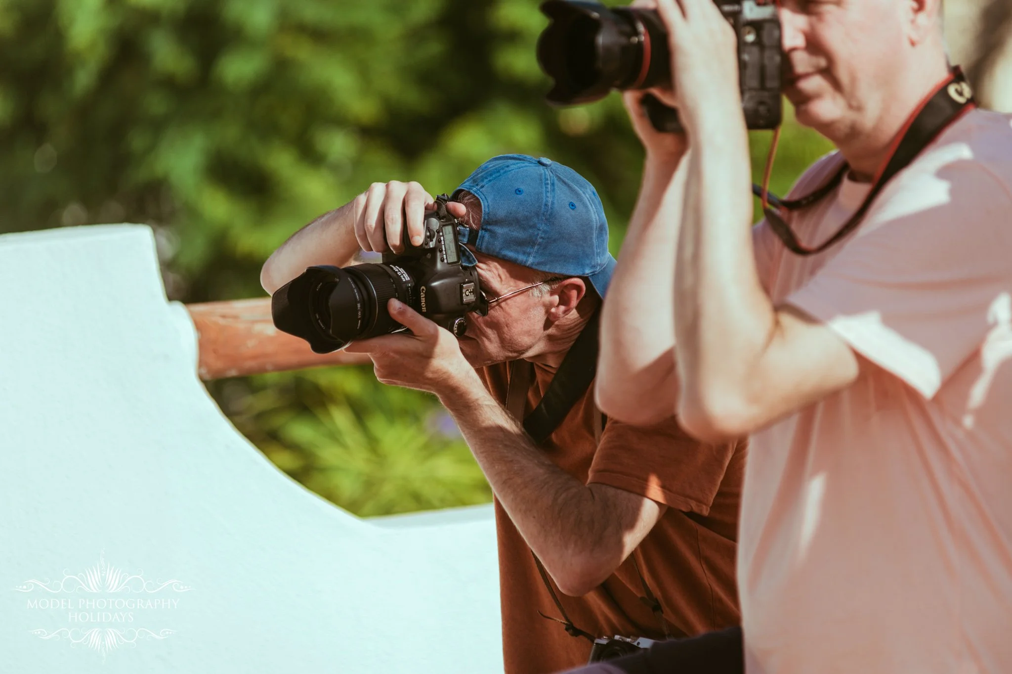 Two photographers, one elderly man wearing a blue cap and brown shirt and another person in a light pink shirt, are taking pictures outdoors using professional cameras. The background is blurred with green foliage, indicating they are in a nature set