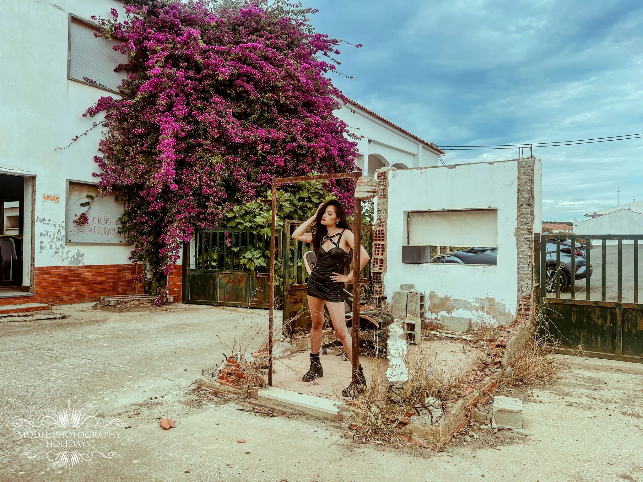 A woman in a black dress and boots standing outdoors in front of a partially constructed or demolished building with a large pink flowering bush behind her.
