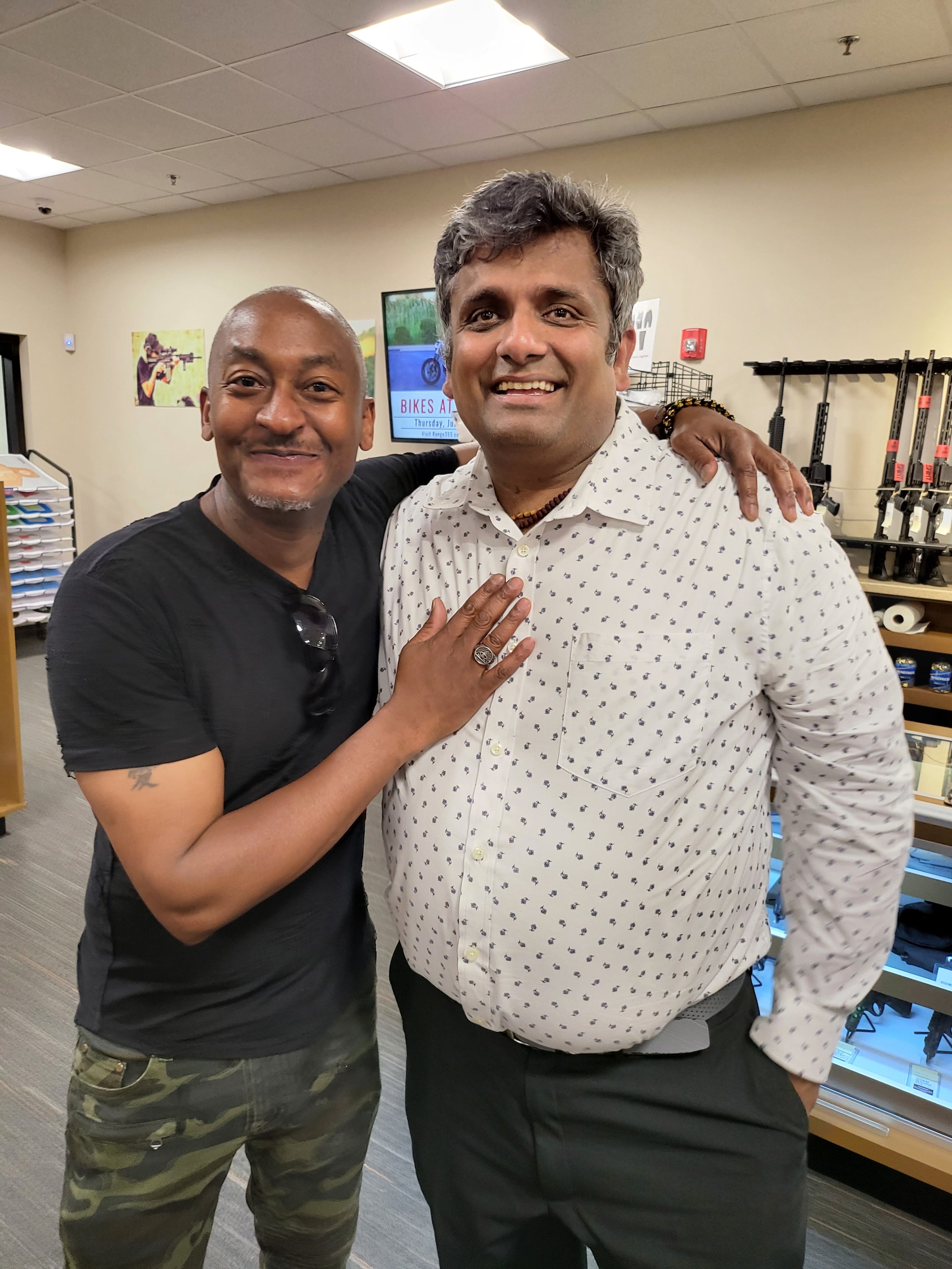 Two men smiling and standing close together indoors, one with his arm around the other, in a room with shelves and a TV screen in the background.