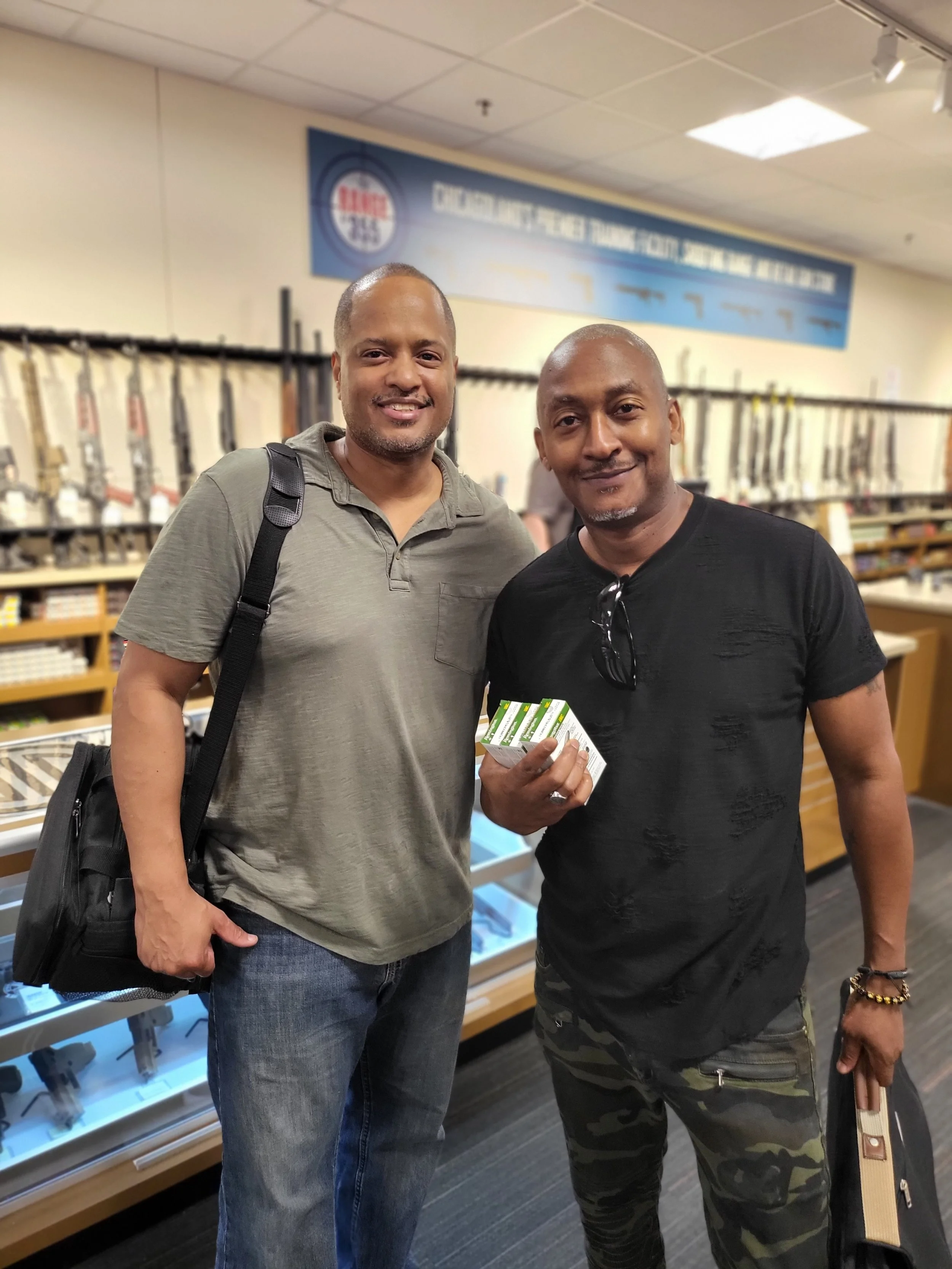 Two men standing in a gun store, smiling at the camera. One is wearing a gray polo shirt and carrying a black shoulder bag, the other is wearing a black t-shirt with glasses hanging from the collar and camouflage pants, holding a boxed item. Seen beh
