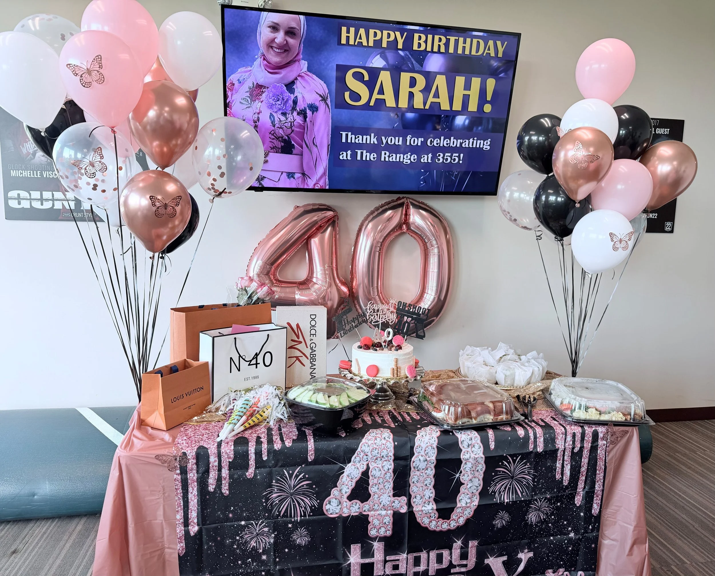 A birthday celebration setup with pink, black, and white balloons, some with butterfly designs, grouped on either side of a decorated table. The table has a pink tablecloth, various snacks, a cake with pink decorations, and birthday-themed items. Beh