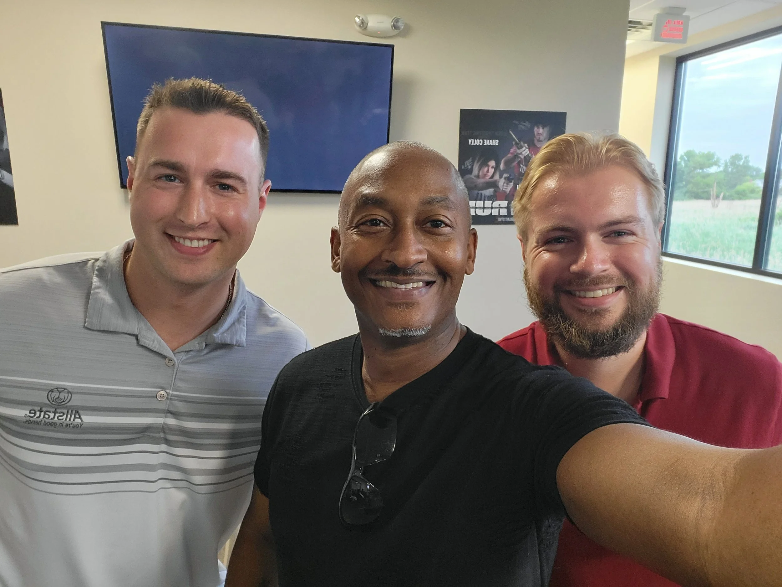 Three men are taking a selfie in a room with a large window. The man on the left wears a light gray shirt, the man in the middle is bald with a dark shirt, and the man on the right has blonde hair and a beard, wearing a red shirt. They are smiling at