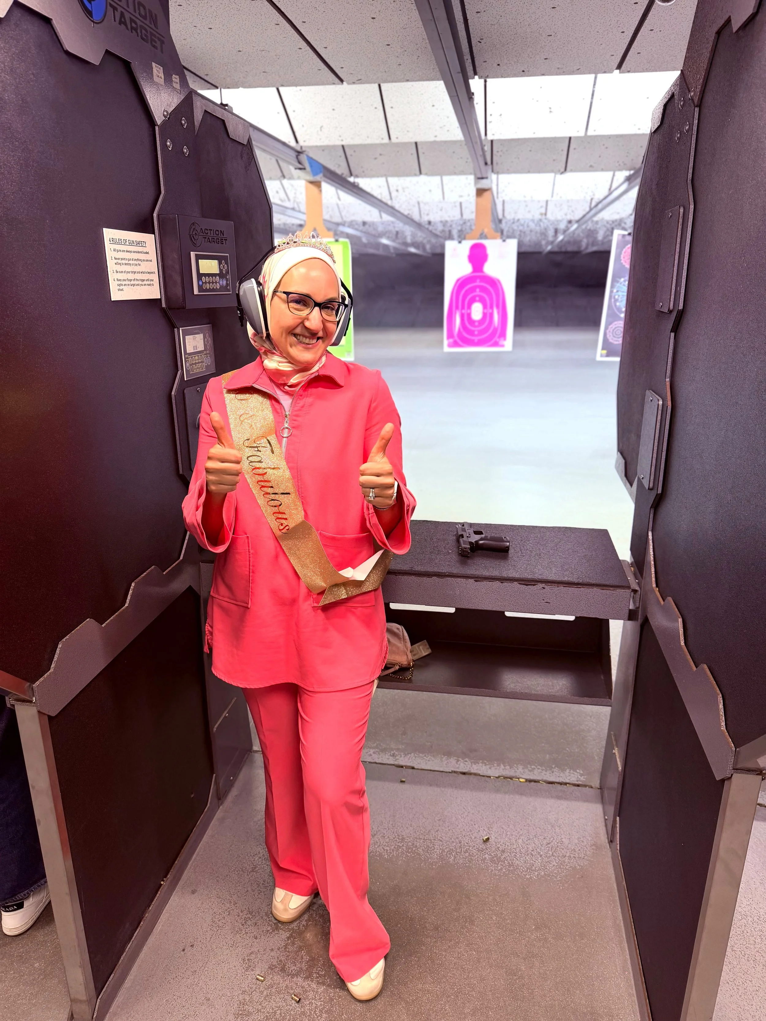 A smiling woman in pink pants, a pink shirt, a decorated headscarf, and glasses, standing in a shooting range or target practice area with gun and target silhouettes in the background.