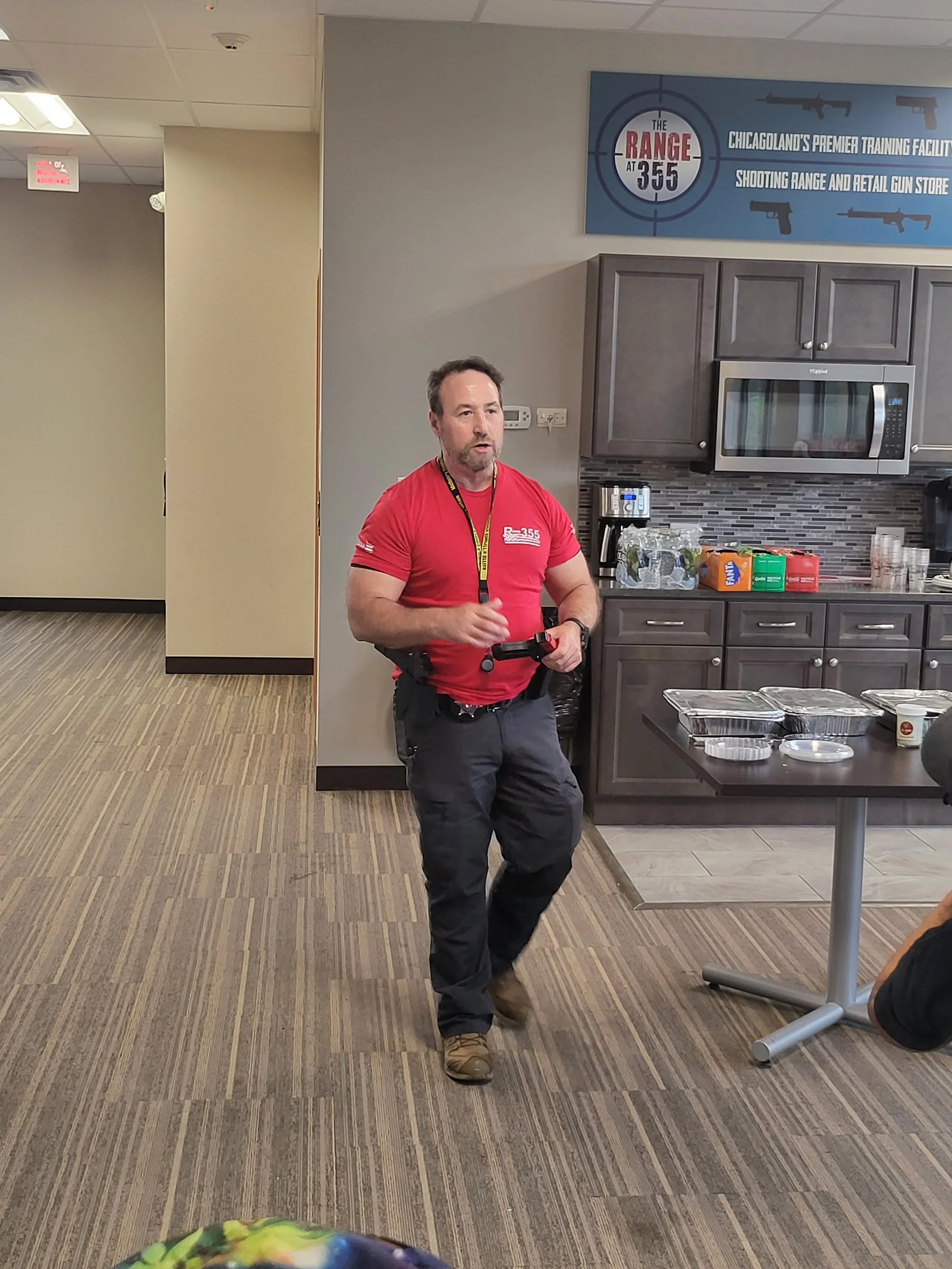 A man with a beard and short hair wearing a red t-shirt and black tactical gear is standing in a room with wooden cabinets, a microwave, and snacks on the counter. The room has gray walls, patterned carpet, and a blue sign with guns and target graphi