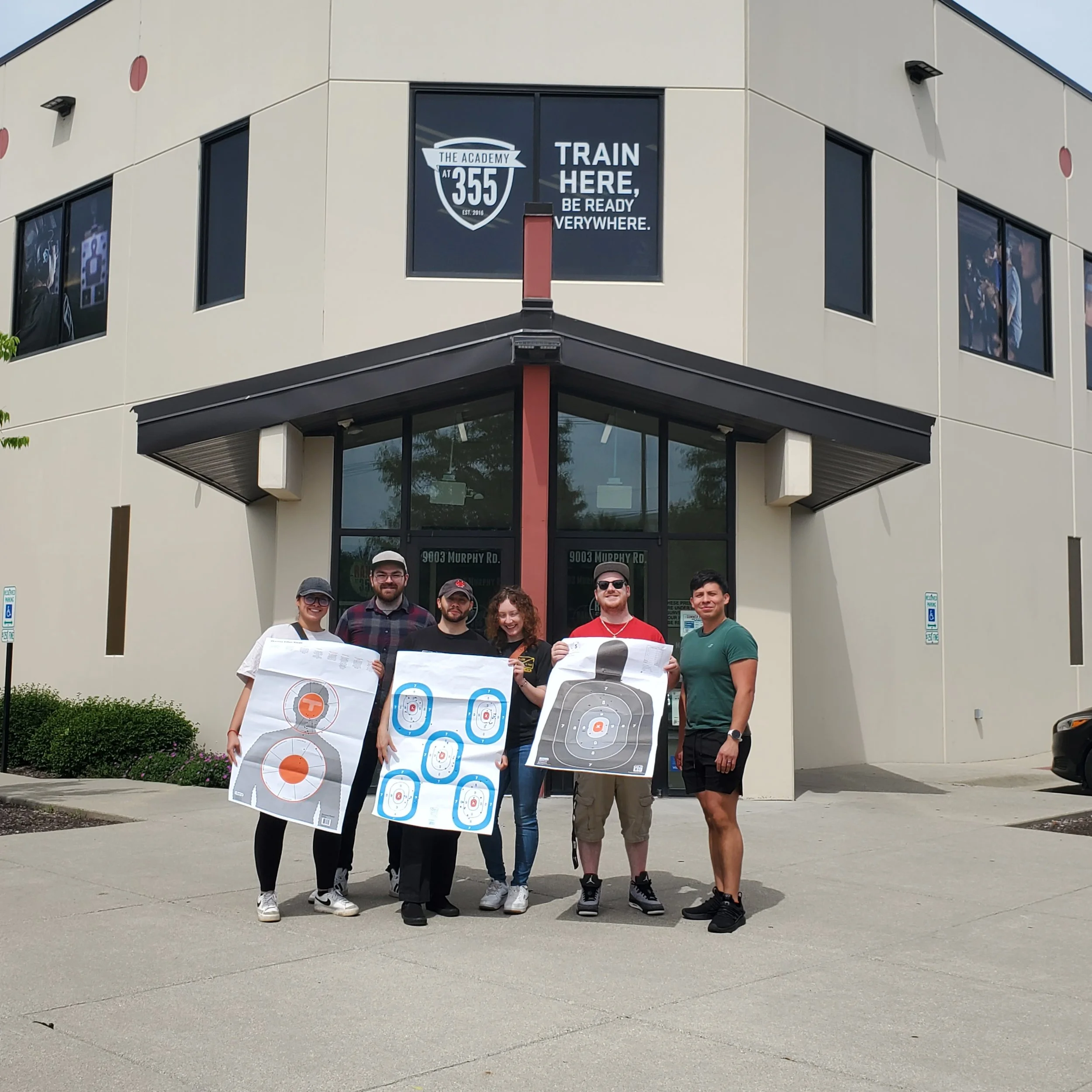 A group of six people standing outside holding shooting target posters in front of a beige building. The building has a sign that reads "The Academy at 355" with a shield logo and the phrase "Train here, be ready everywhere." There is also a digital 